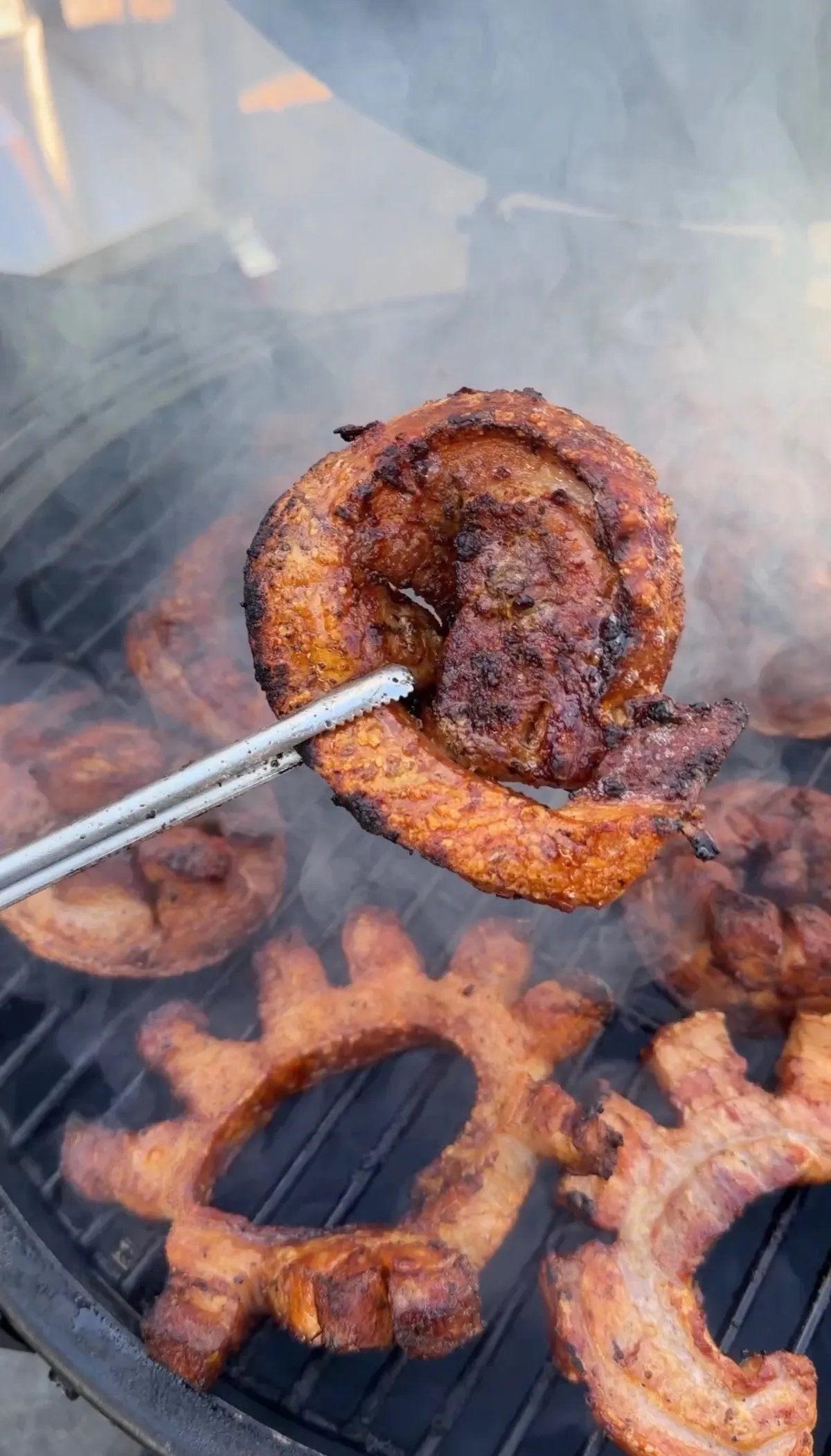 Pork belly pinwheel held with tongs over charcoal grill, skin crisped with ancho chili seasoning, with more pinwheels grilling below