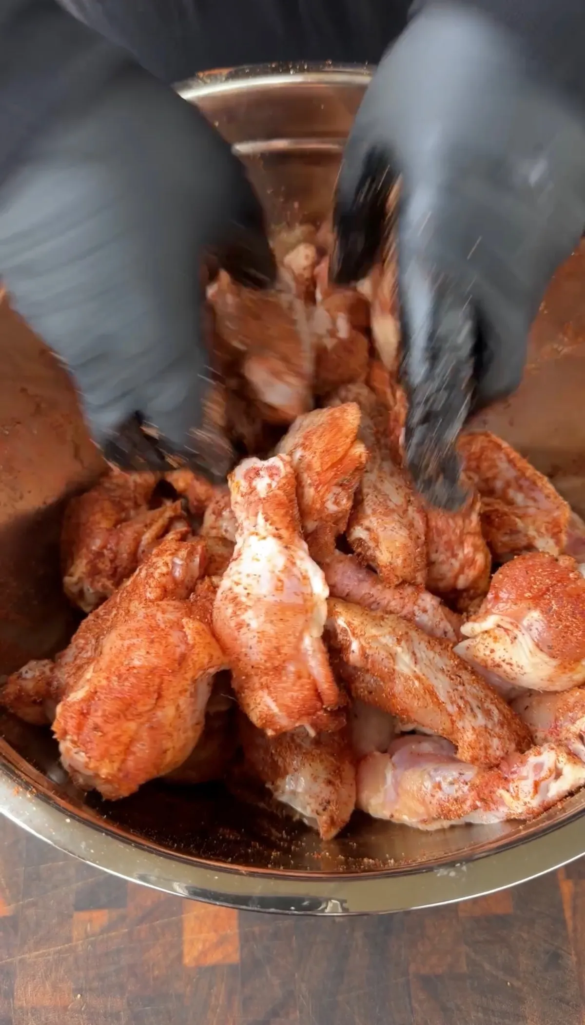 Gloved hands tossing raw chicken wings coated in chipotle seasoning in a stainless steel mixing bowl