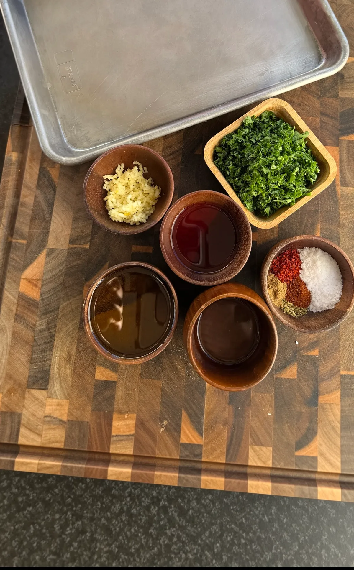 Chimichurri sauce ingredients in wooden bowls: minced garlic, chopped parsley, red wine vinegar, olive oil, and spices on a cutting board