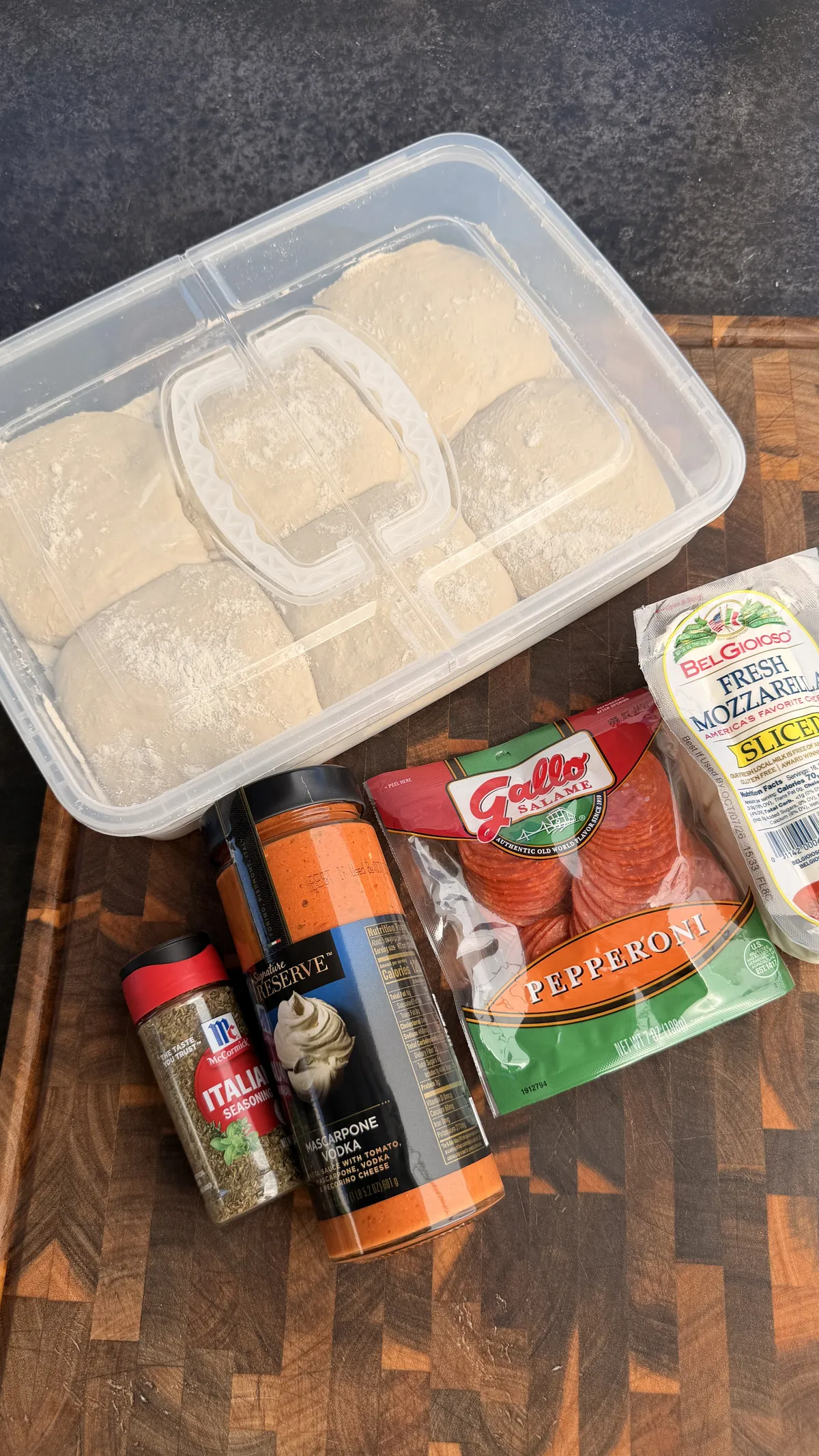 Pizza dough balls in container with pepperoni, fresh mozzarella, Italian seasoning, and pizza sauce on a wooden cutting board