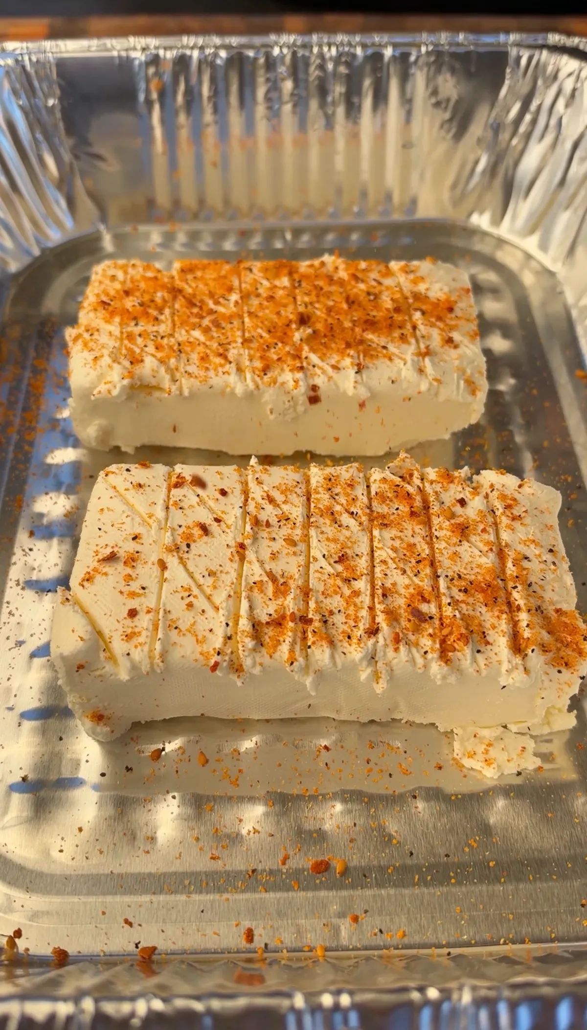 Two 8-oz cream cheese blocks scored in crosshatch pattern and seasoned with seafood seasoning in aluminum tray, ready to smoke