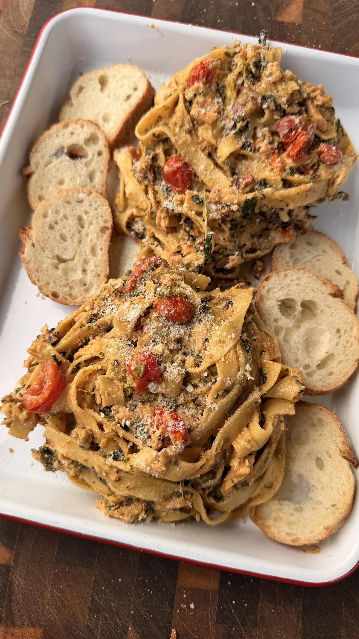 Two servings of creamy pesto salmon fettuccine with cherry tomatoes, spinach, and Parmesan on a white tray with toasted bread slices