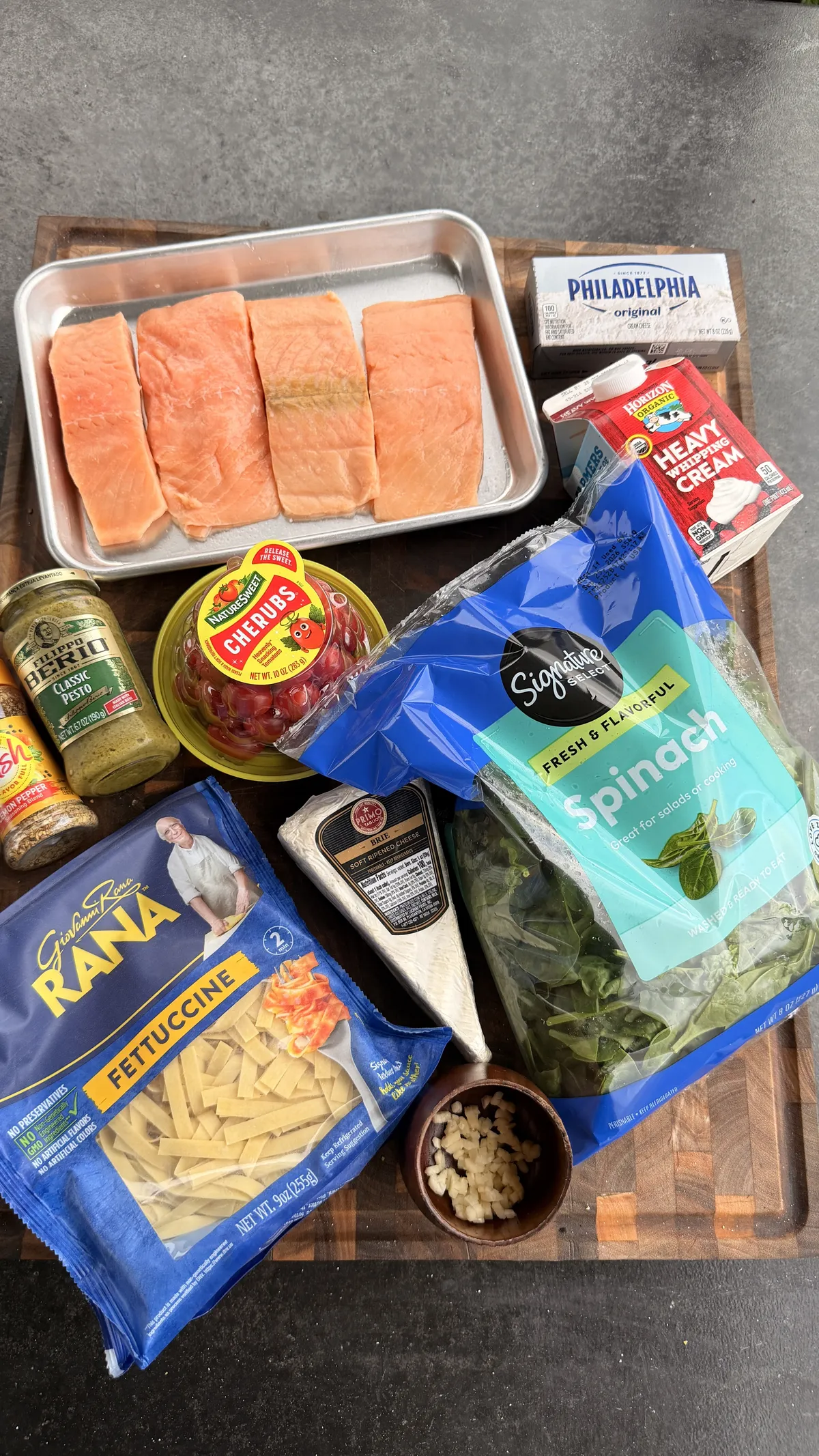 Flat lay of ingredients for creamy pesto salmon pasta: salmon portions, Brie, cream cheese, heavy cream, pesto, fettuccine, spinach, cherry tomatoes, and garlic