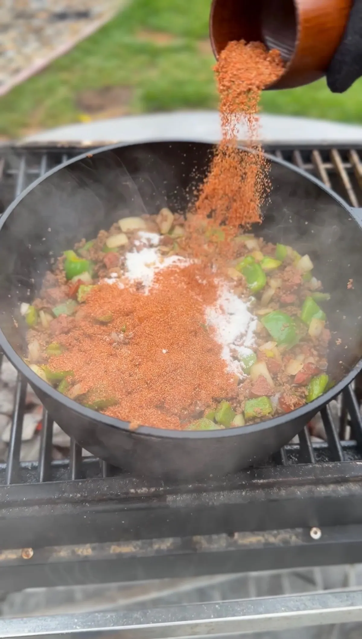 Creole seasoning and flour being poured over sautéed green bell pepper and onion in a Dutch oven on a grill