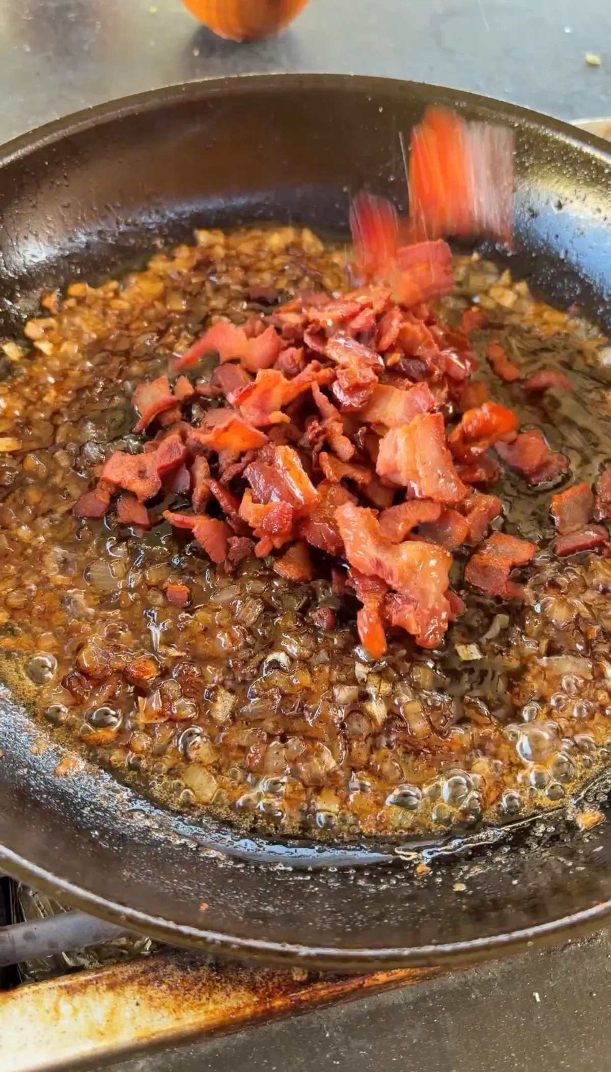 Crispy chopped bacon being added to caramelized onion and bourbon glaze in a skillet for homemade bacon jam