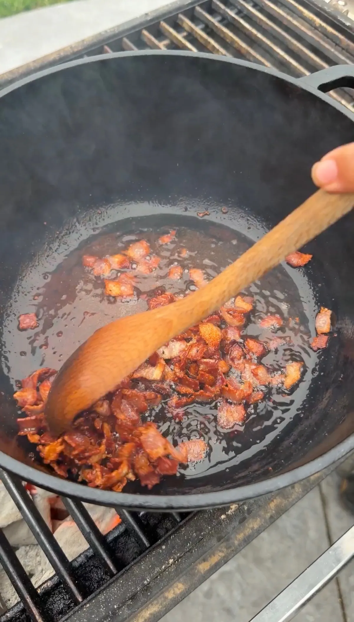 Crispy bacon pieces rendering in a large cast iron Dutch oven on a grill, stirred with a wooden spoon