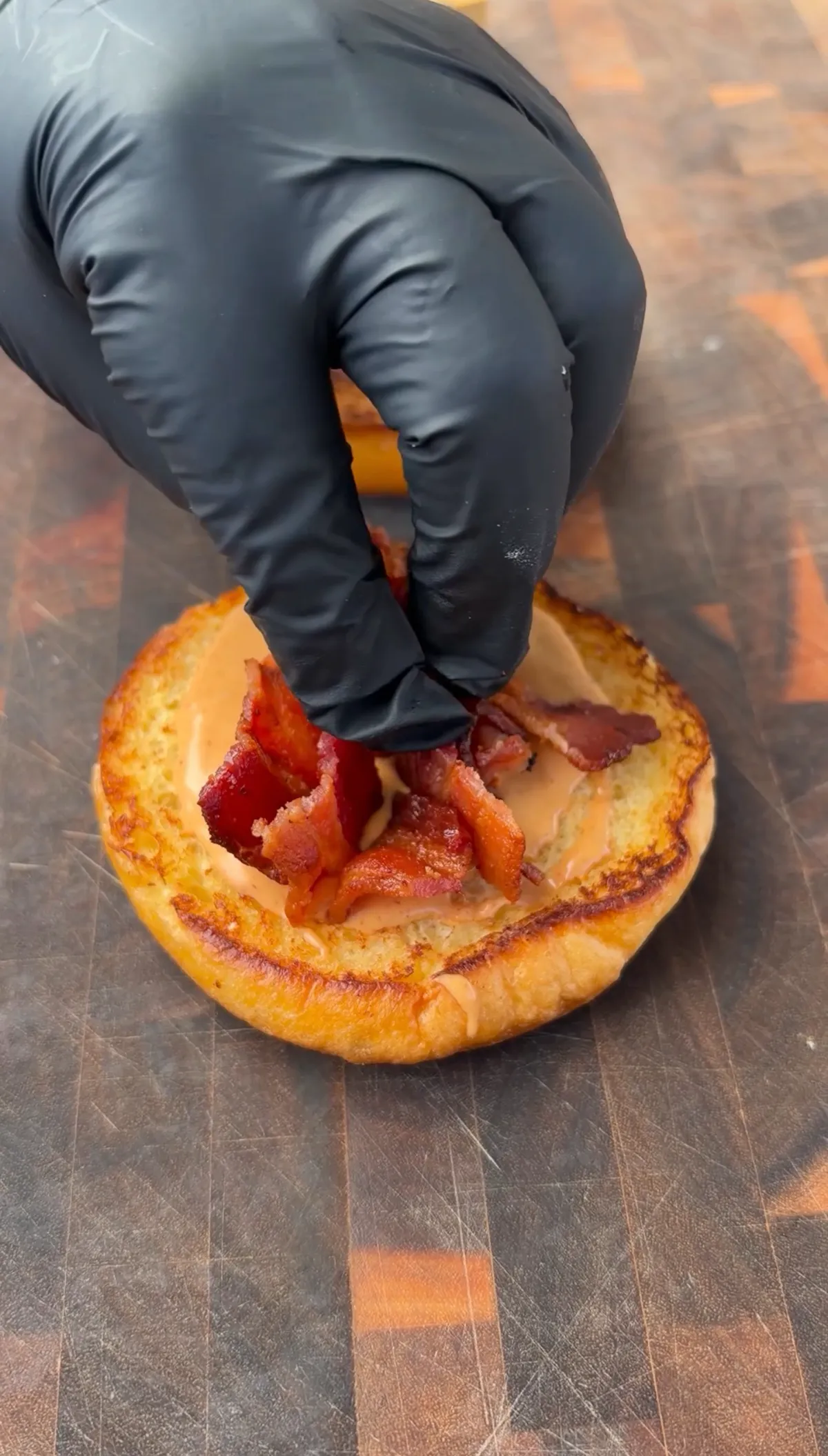 Gloved hand placing crispy chopped bacon on a toasted buttered brioche bun spread with chipotle mayo