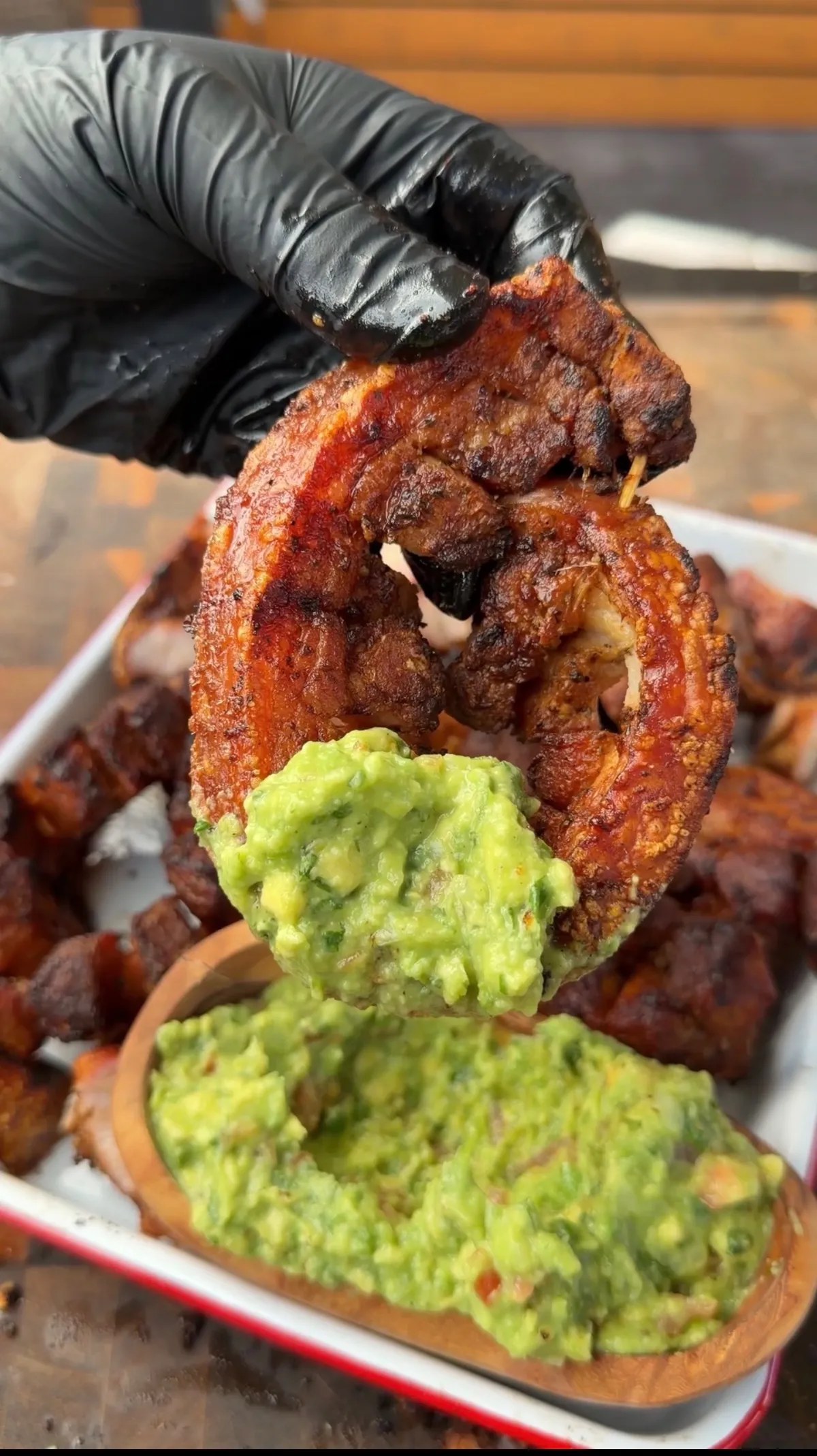 Grilled chicharrón pinwheel held over fresh guacamole in a molcajete, with charcoal-cooked pork belly in tray behind