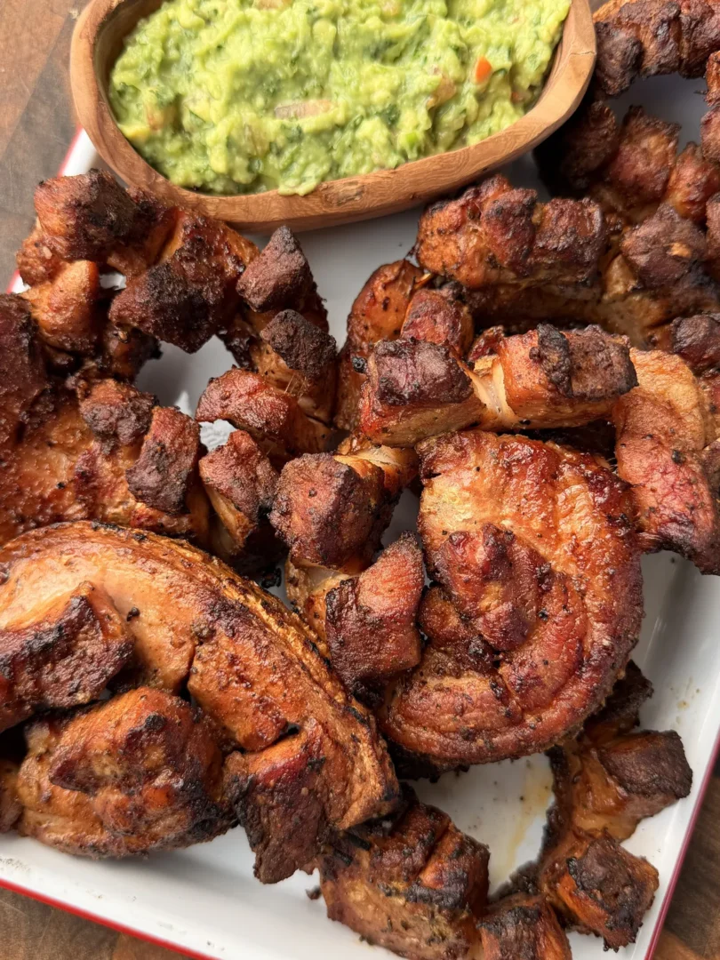Grilled chicharrón pinwheels with crispy skin on a white platter served with fresh guacamole in a wooden bowl