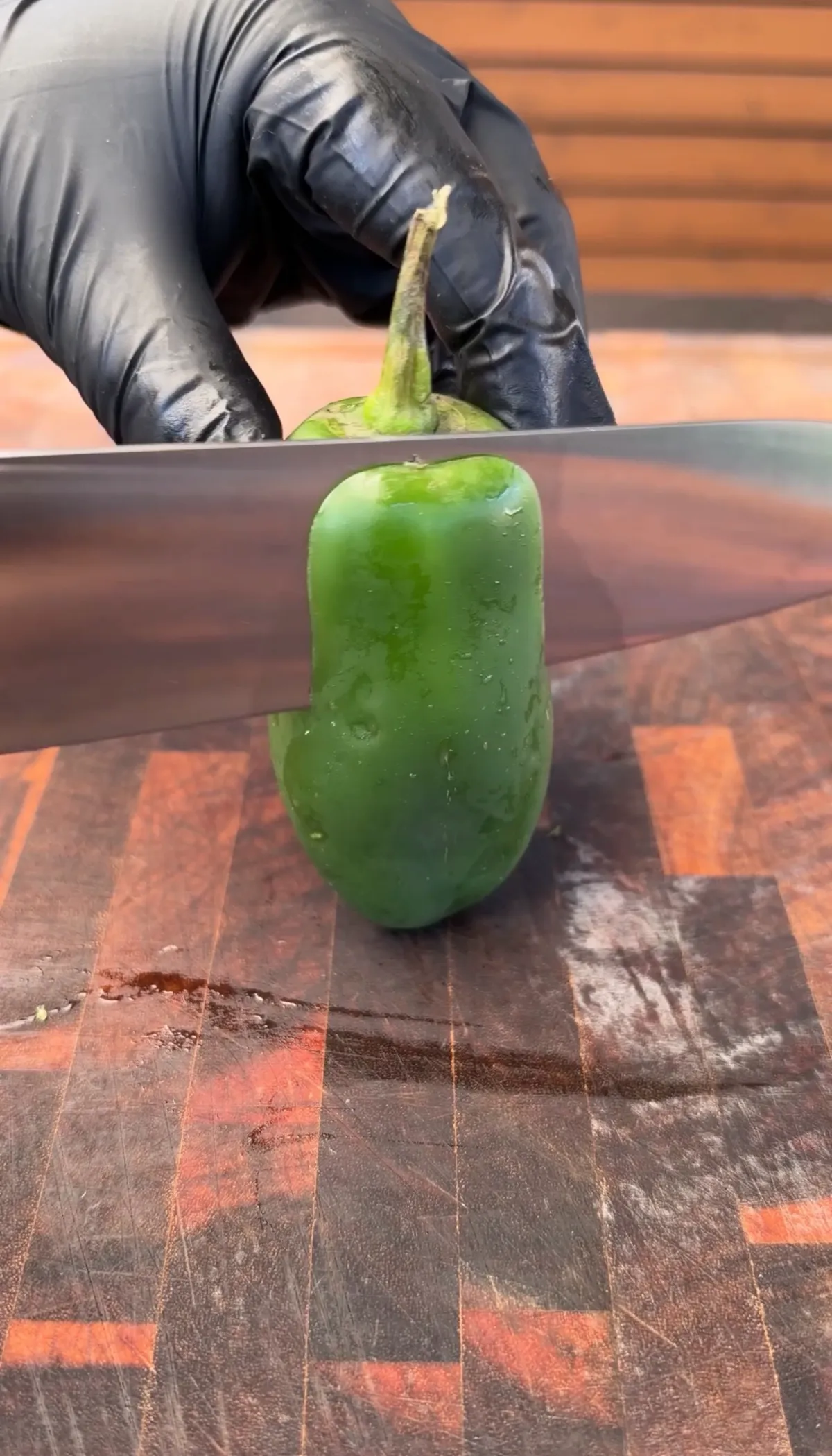 Black-gloved hand using a sharp knife to cut the top quarter off a large jalapeño standing upright on a wood cutting board