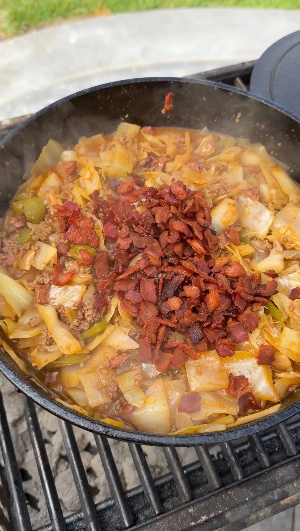 Crispy bacon added to dirty cabbage with ground chuck, kielbasa, bell peppers, and onions simmering in a Dutch oven on a grill