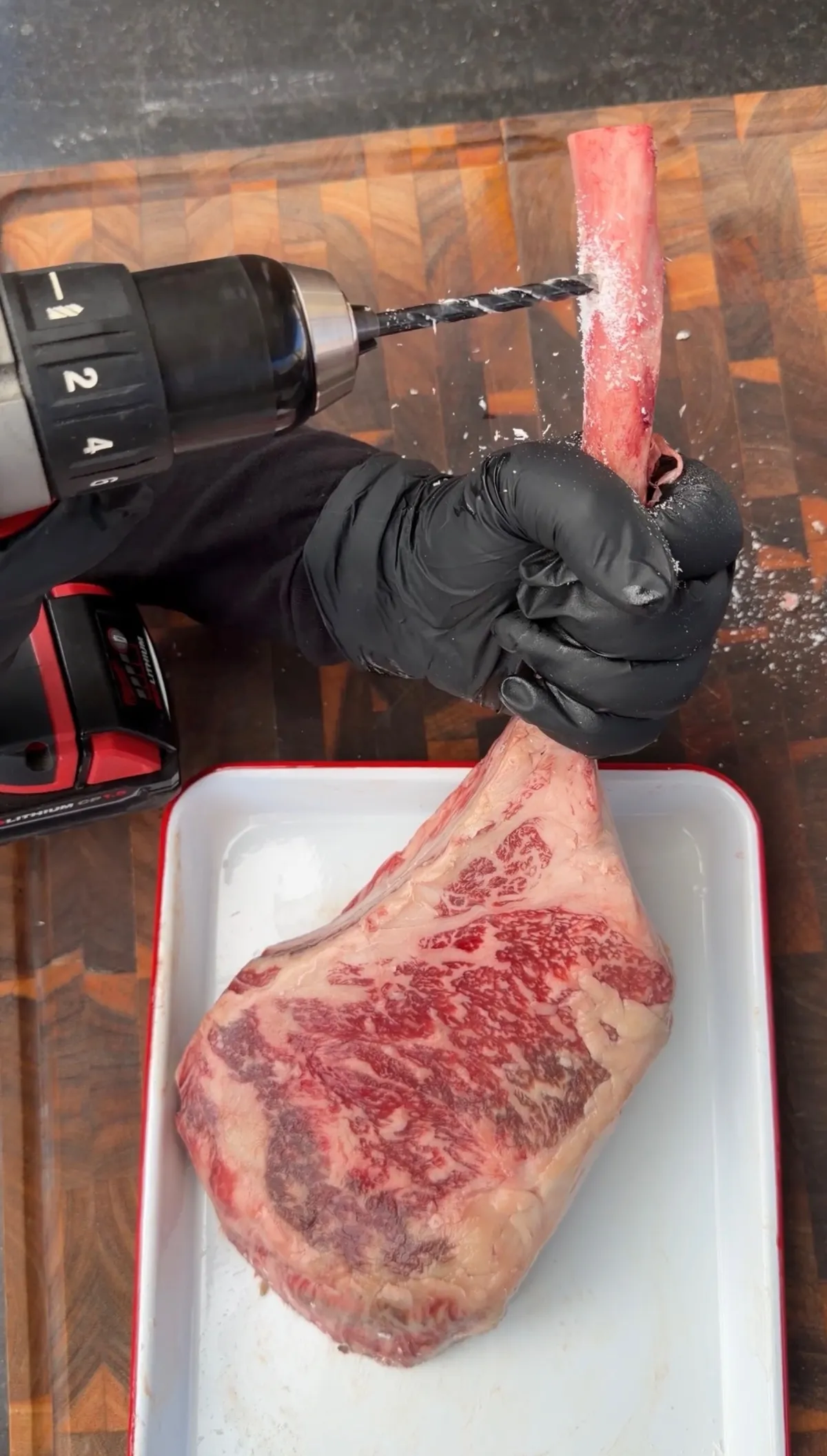 Power drill creating hole through tomahawk steak bone for smoker hanging setup on wood cutting board