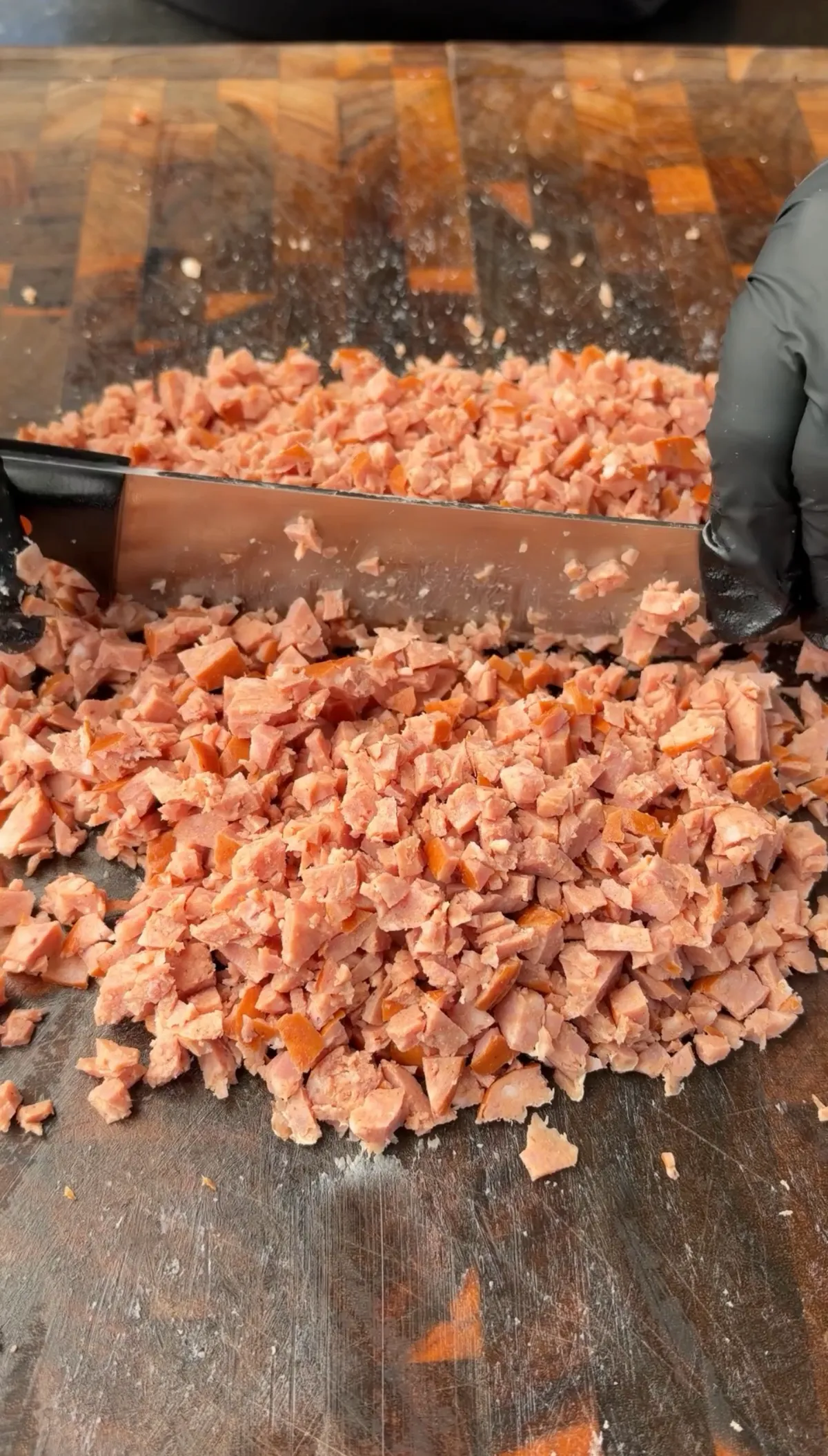 Finely diced kielbasa sausage being chopped on a wooden cutting board with a bench scraper, gloved hands visible