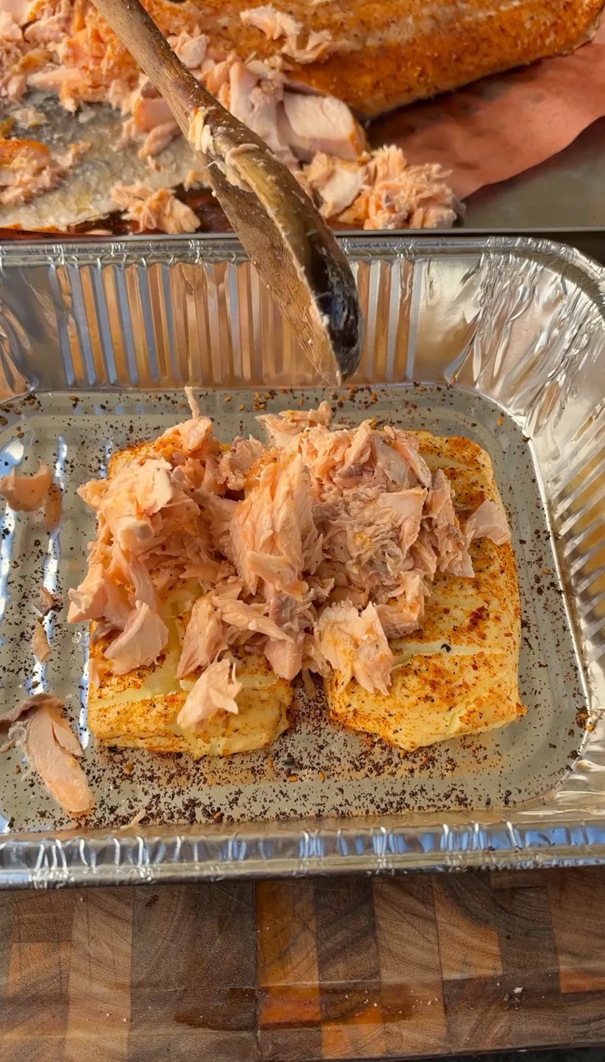 Flaked smoked salmon being added over two scored, seasoned cream cheese blocks in a foil tray