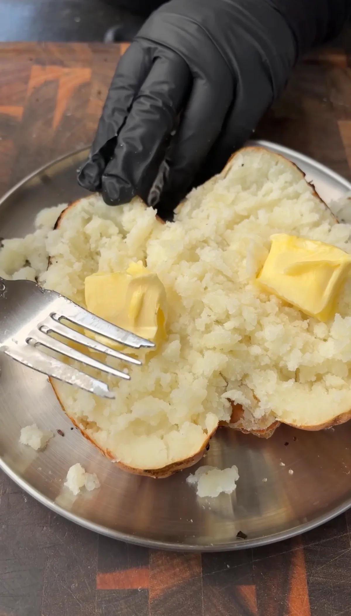 Gloved hand fluffing russet potato half with fork, topped with two pats of butter on a steel plate
