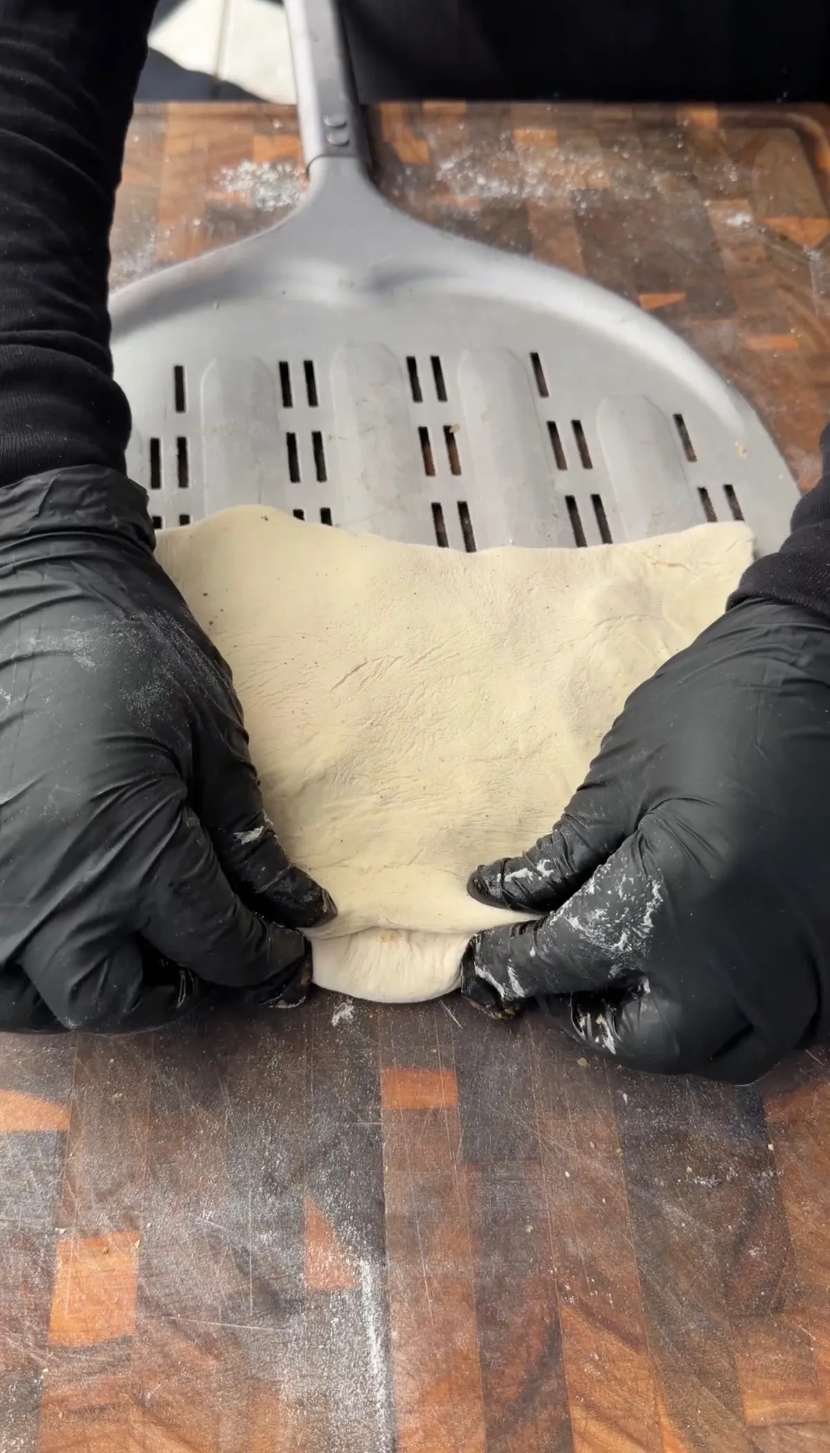 Hands in black gloves folding and crimping pizza dough edges to seal a classic pepperoni calzone on a floured wooden board