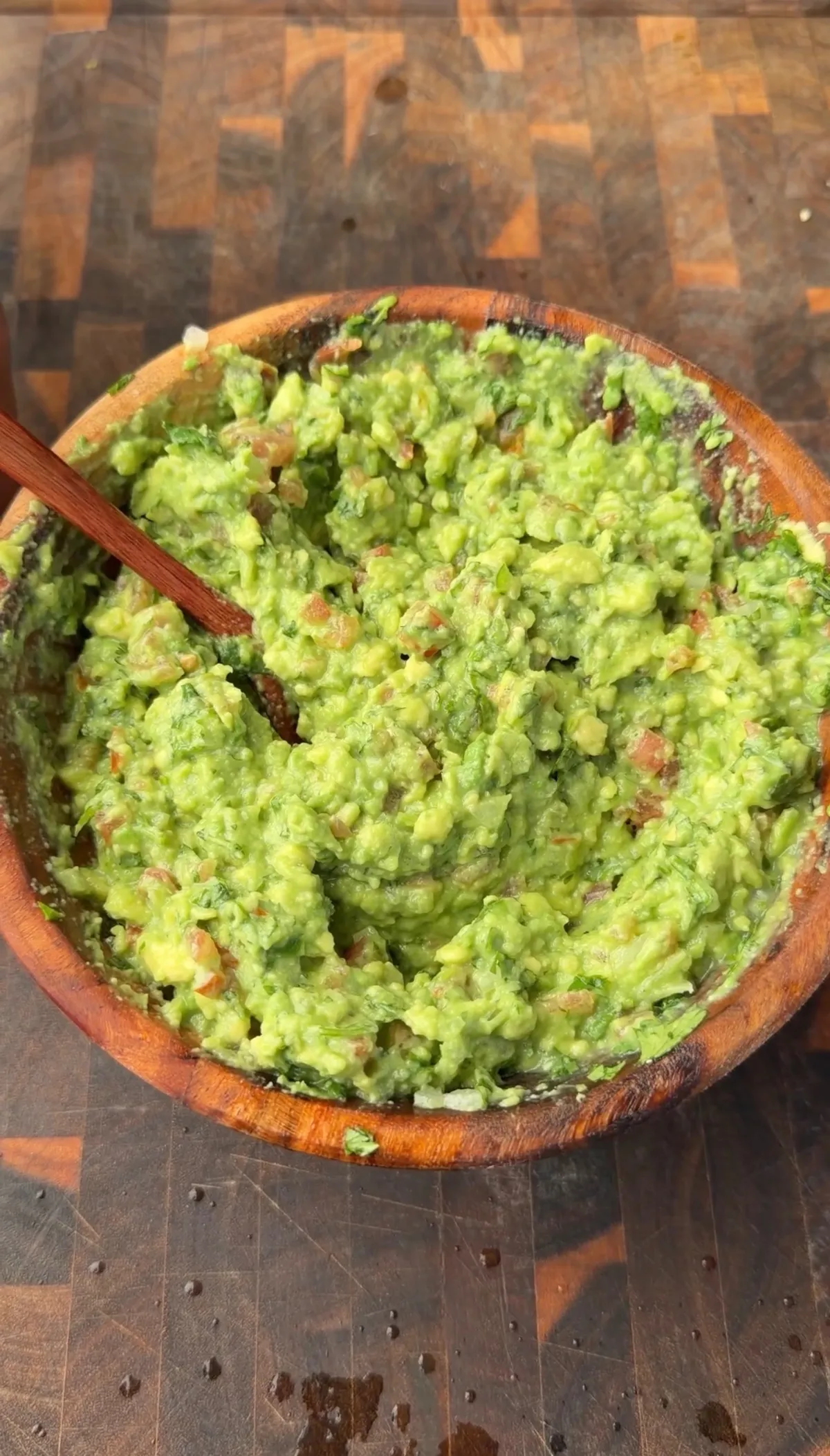 Fresh guacamole with diced tomato, yellow onion, Serrano pepper, and cilantro in a wooden bowl with a wooden spoon