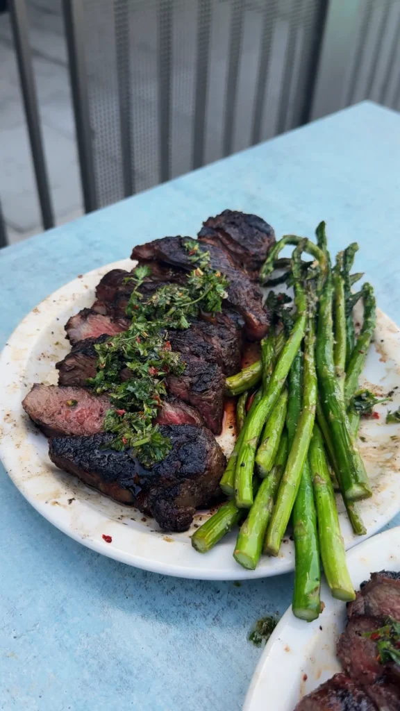 Sliced grilled New York strip steak topped with chimichurri beside pan-roasted asparagus on a white plate