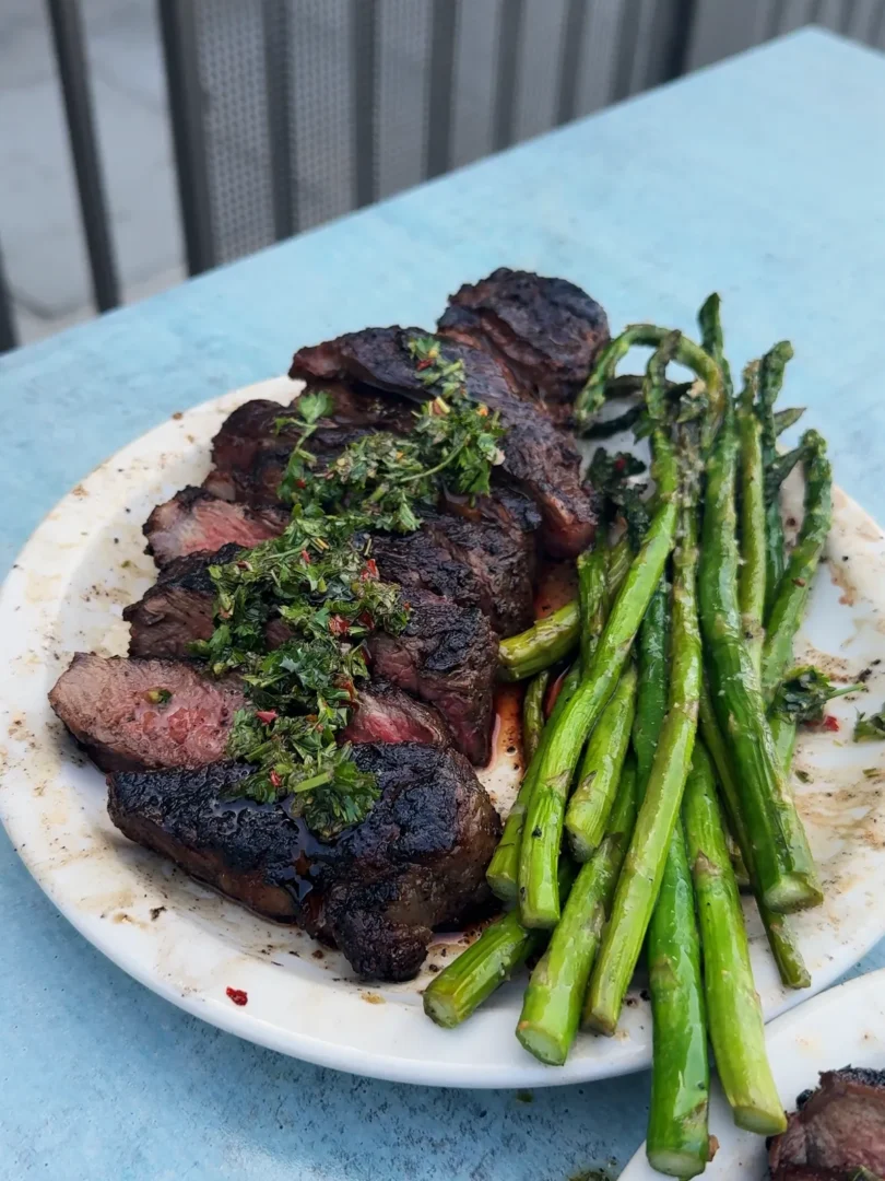 Sliced grilled New York strip steak topped with chimichurri beside pan-roasted asparagus on a white plate