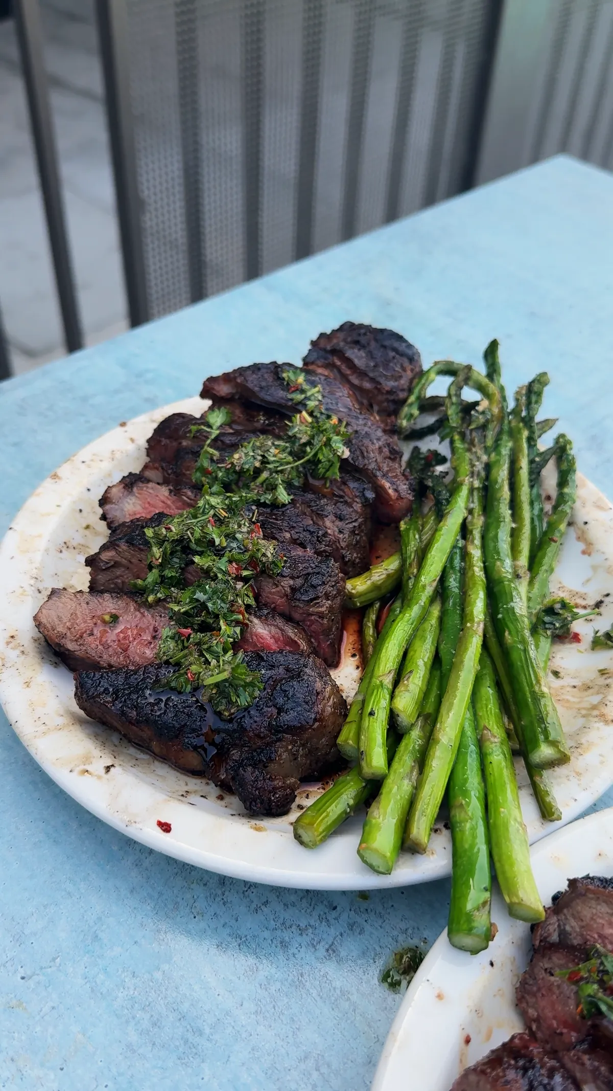 Sliced grilled New York strip steak topped with chimichurri beside pan-roasted asparagus on a white plate