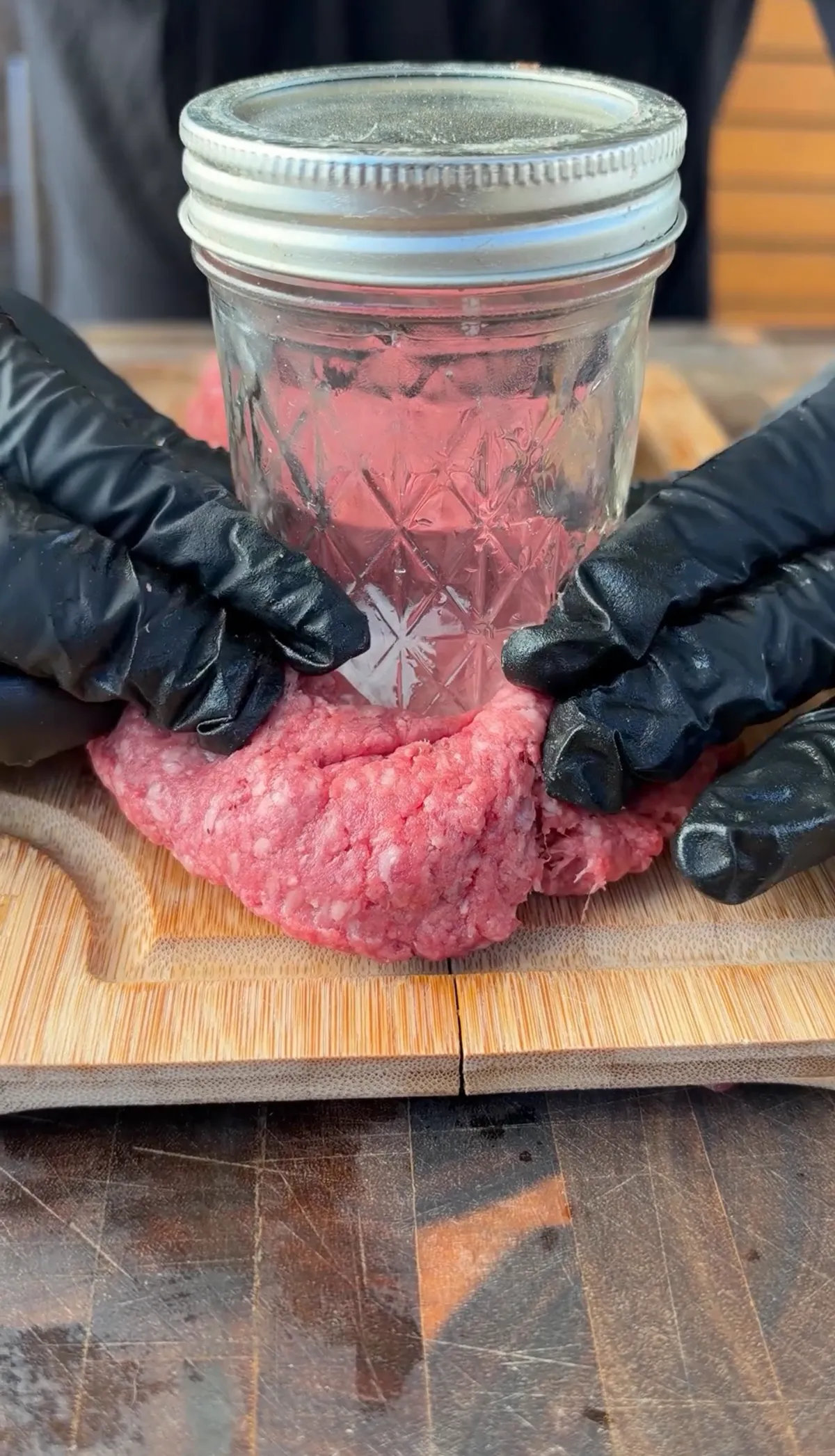 Gloved hands pressing a Mason jar into an 85/15 ground beef patty on a cutting board to form a stuffed burger pocket