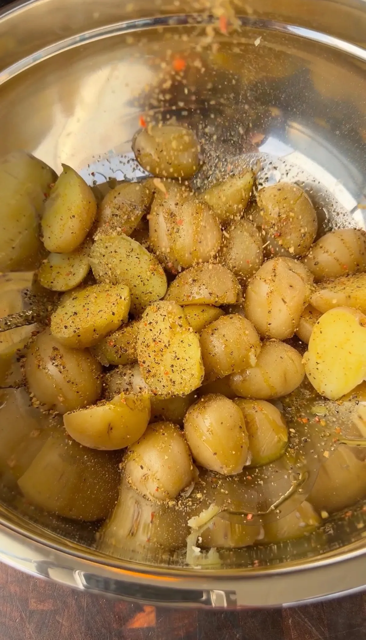 Halved baby potatoes tossed with avocado oil and lemon pepper seasoning in a stainless steel mixing bowl