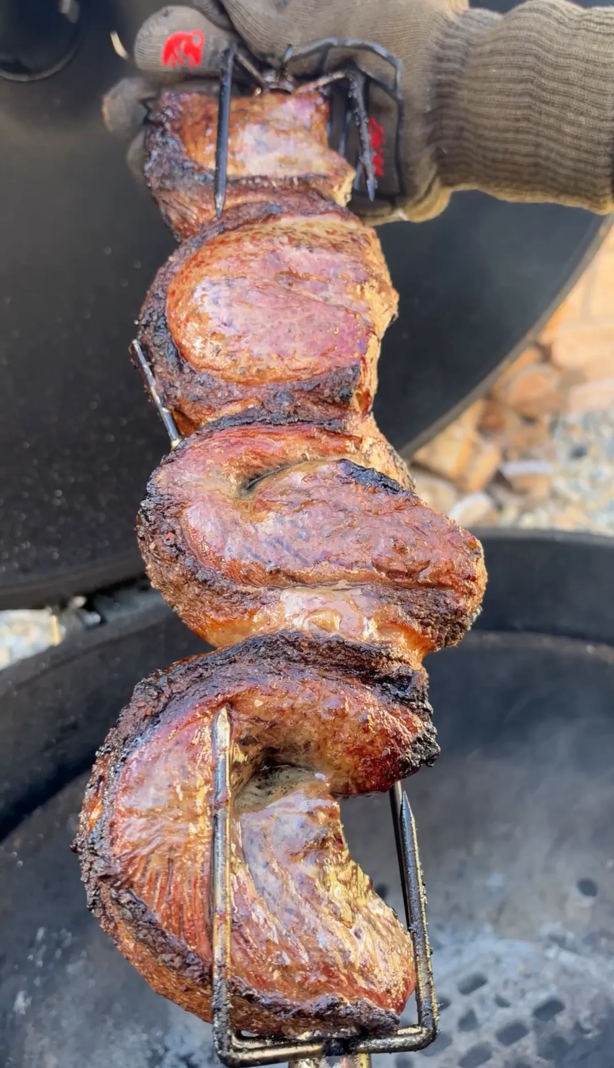 Whole picanha steaks folded C-shape and threaded on a rotisserie spit, fat cap out, over a charcoal grill