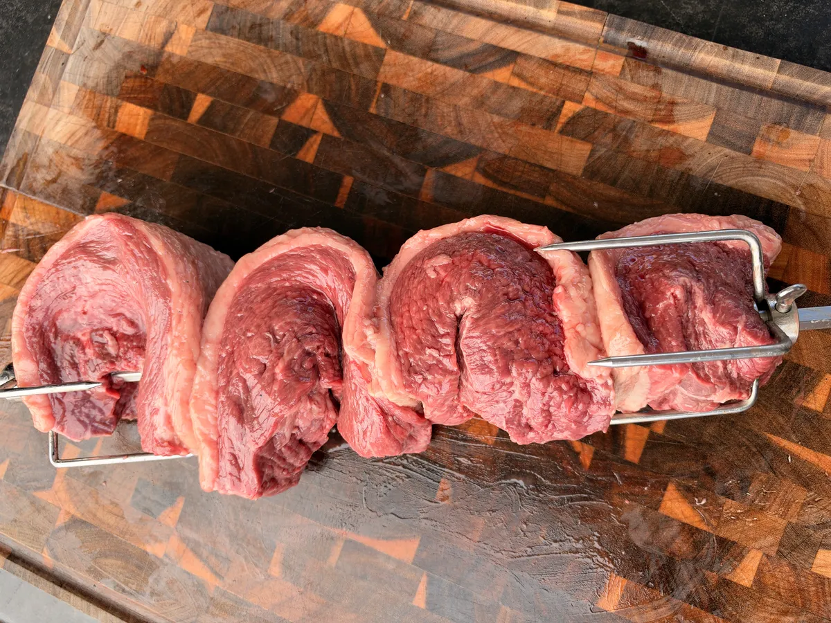Four picanha steaks folded Brazilian churrasco-style into a C shape, threaded on a rotisserie spit on a wooden cutting board