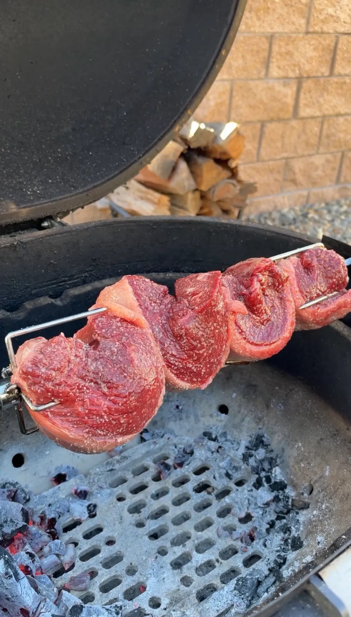 Raw picanha steaks folded Brazilian churrasco-style into C shapes, threaded on a rotisserie spit over hot charcoal