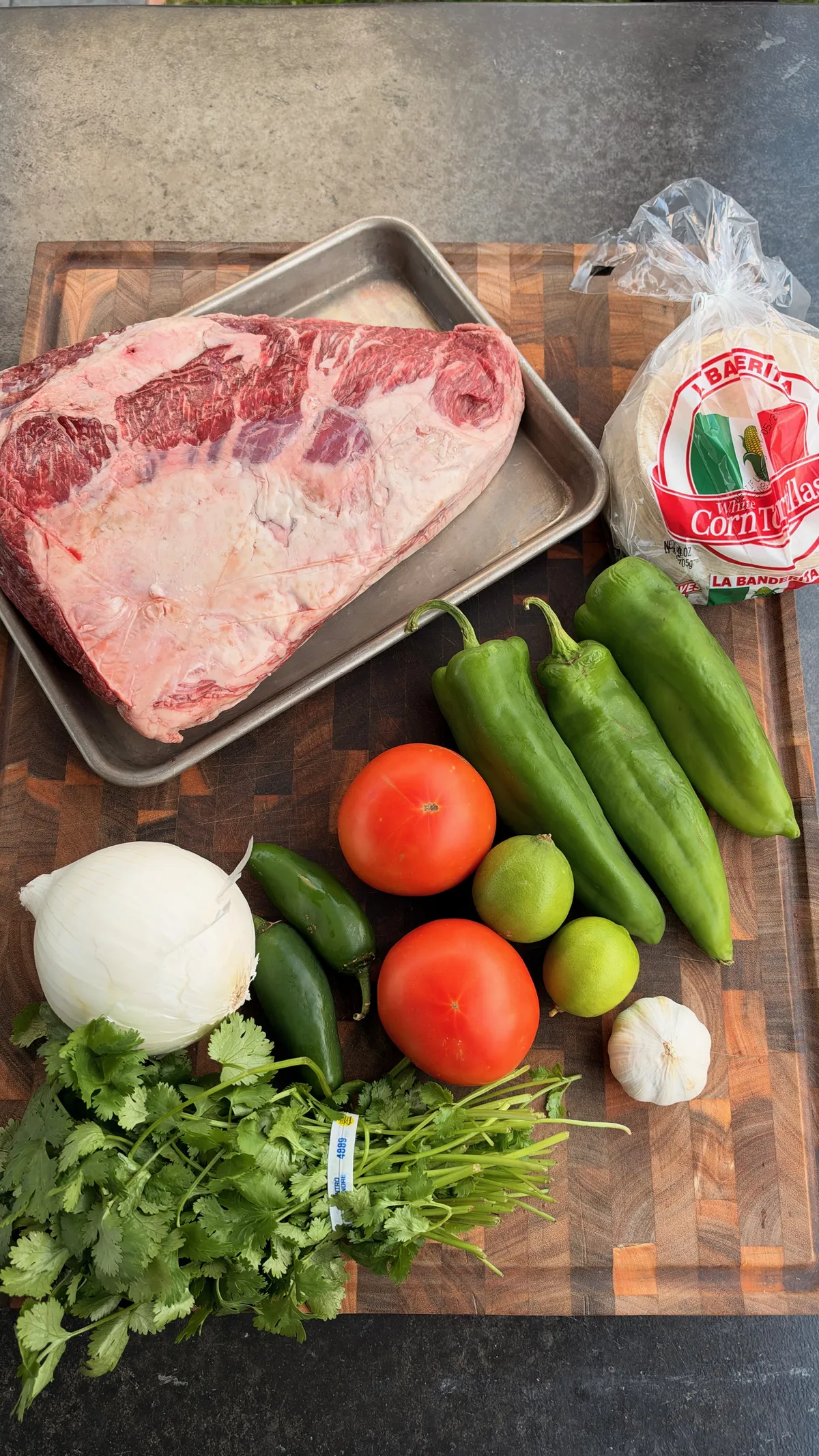 Whole picanha, Hatch green chiles, Roma tomatoes, jalapeño, white onion, cilantro, limes, garlic, and corn tortillas on a cutting board