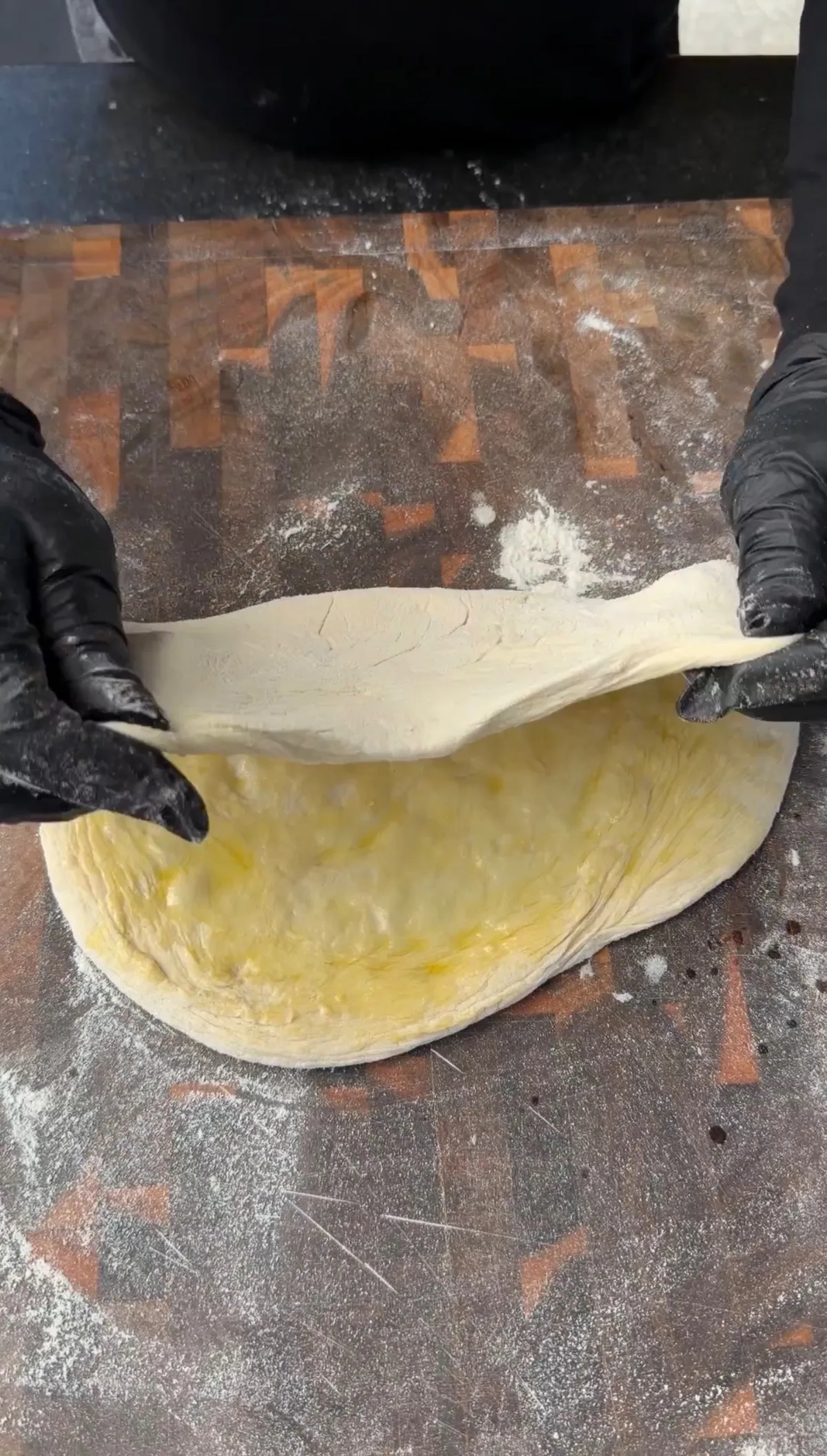 Hands in black gloves folding olive oil-brushed pizza dough in half on a floured surface to make a pizza sandwich