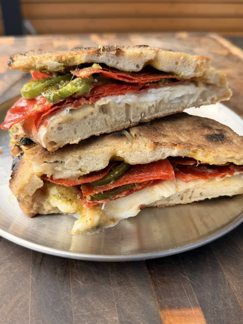 Pizza sandwich cut in half showing burrata, crispy pepperoni, charred jalapeños, hot honey, and pesto on a metal plate