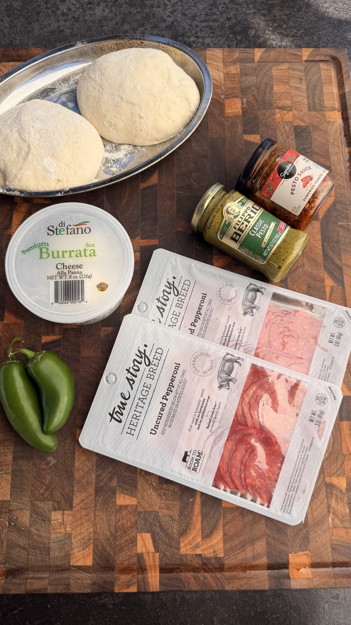 Pizza sandwich ingredients on a wood board: pizza dough balls, burrata, uncured pepperoni, classic pesto, and jalapeños