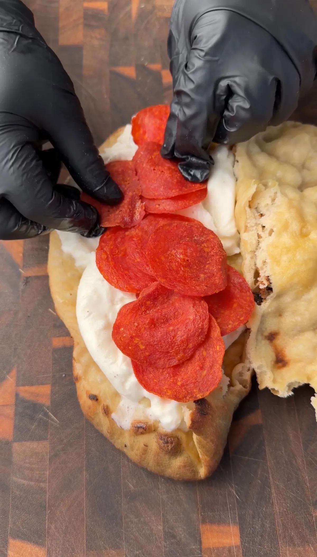 Hands in black gloves layering crispy pepperoni over burrata on an open pizza sandwich on a wooden board
