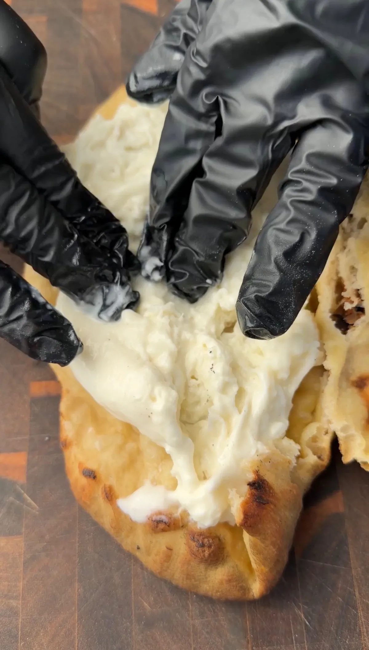 Gloved hands pulling open a golden pizza sandwich revealing creamy burrata cheese on a wooden board