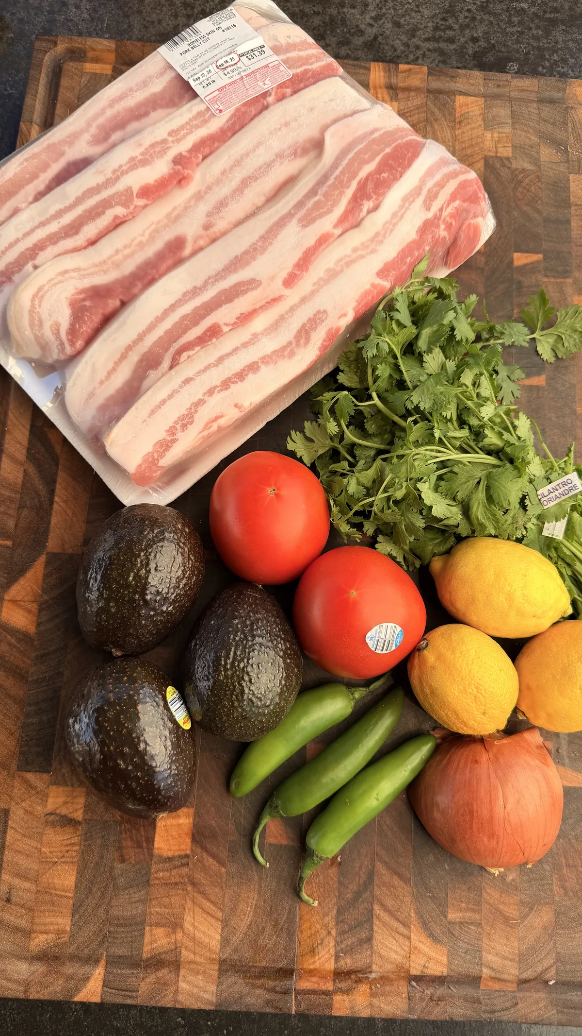 Raw pork belly, avocados, tomatoes, Serrano peppers, cilantro, yellow onion, and lemons on a wooden cutting board