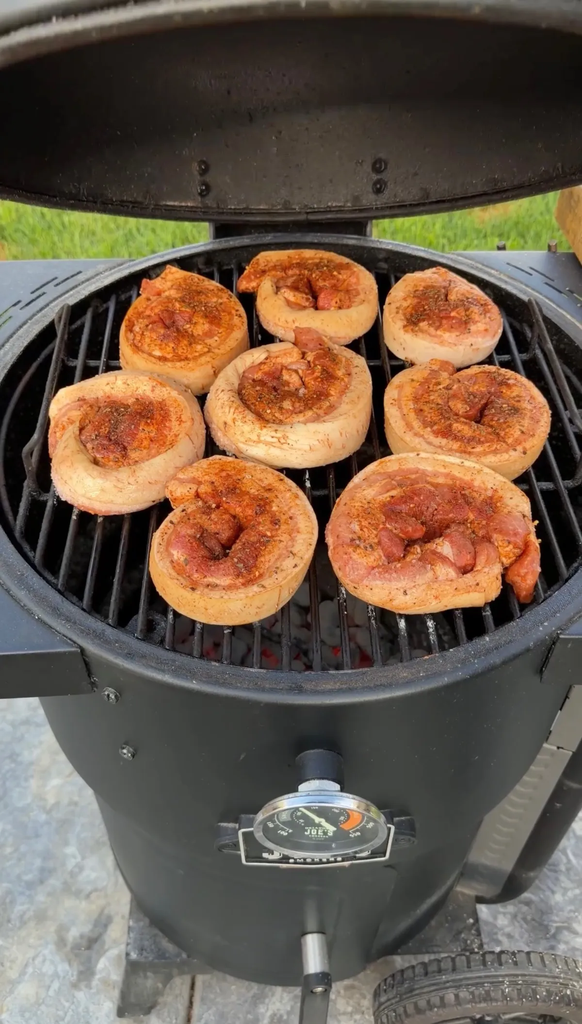 Eight seasoned pork belly pinwheels with ancho chili seasoning cooking over direct charcoal heat on a barrel grill