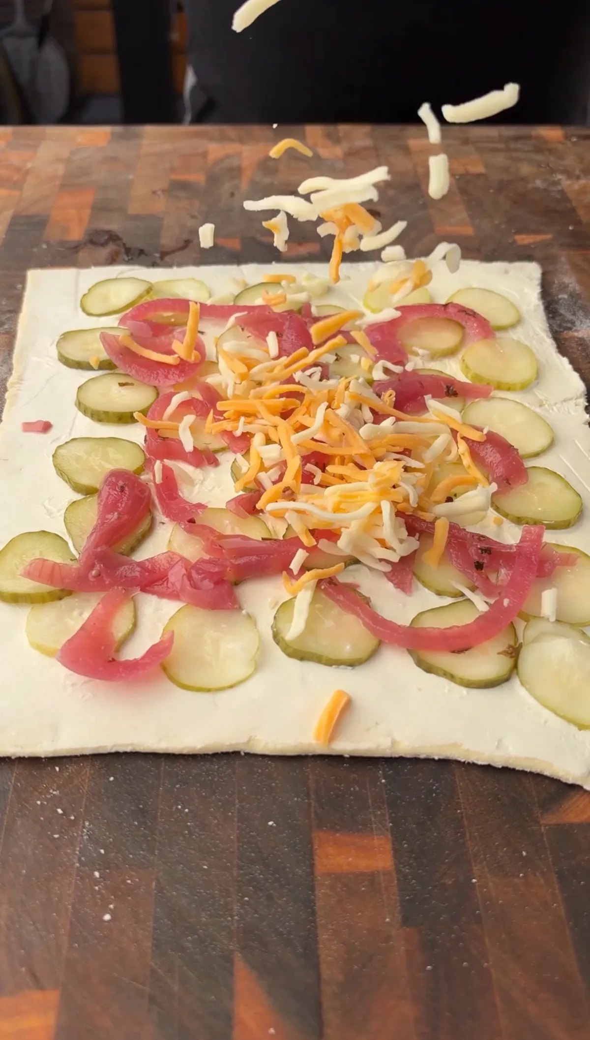 Puff pastry sheet topped with sliced pickles, pickled red onions, and shredded cheddar cheese being added mid-air