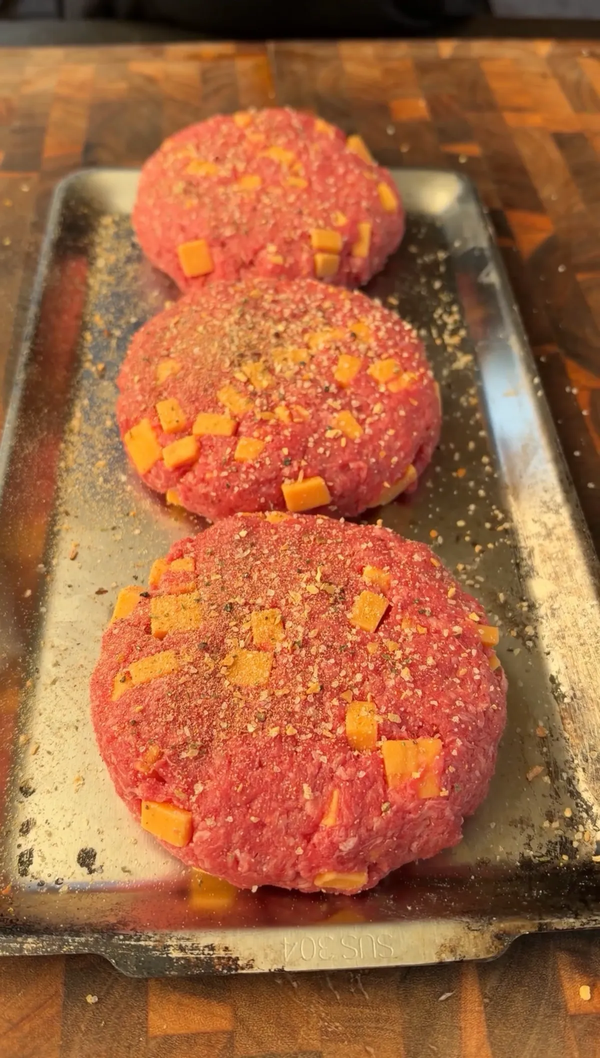 Three raw 85/15 ground beef patties stuffed with cubed cheddar cheese, seasoned with salt, pepper, and garlic powder on a metal tray