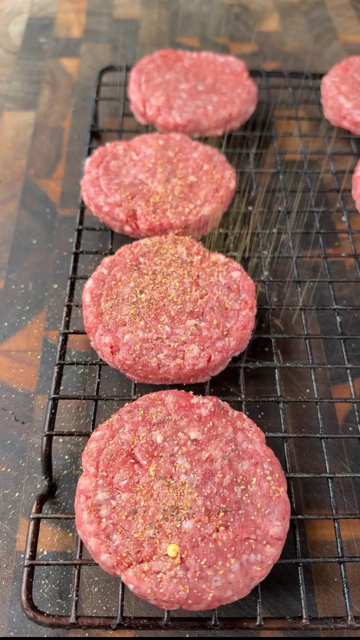 Eight raw 85/15 ground beef slider patties seasoned with Bear Mountain Barbecue Tailgating Blend on a wire rack
