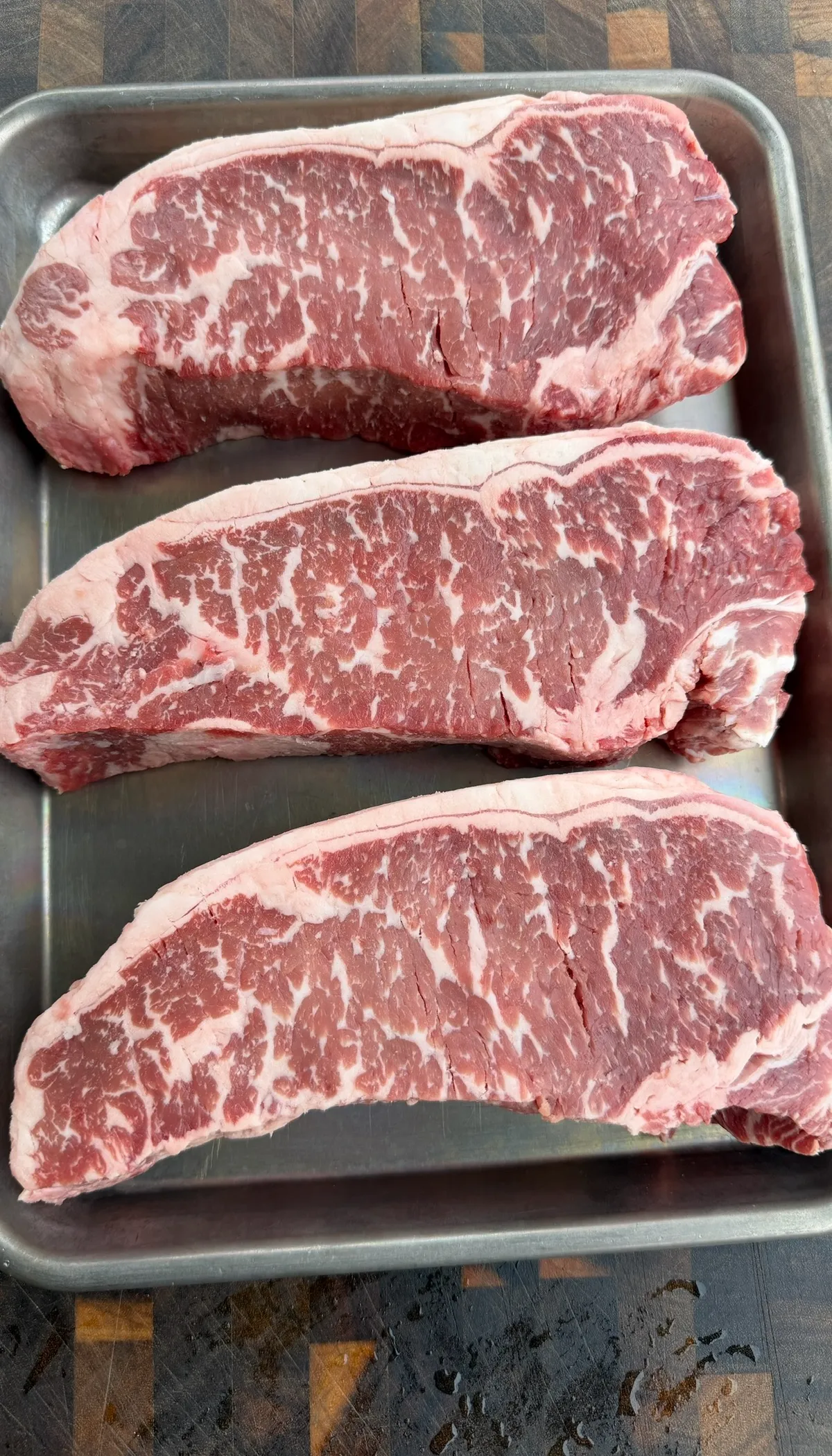 Three raw New York strip steaks with heavy marbling on a stainless steel tray, ready to season and grill
