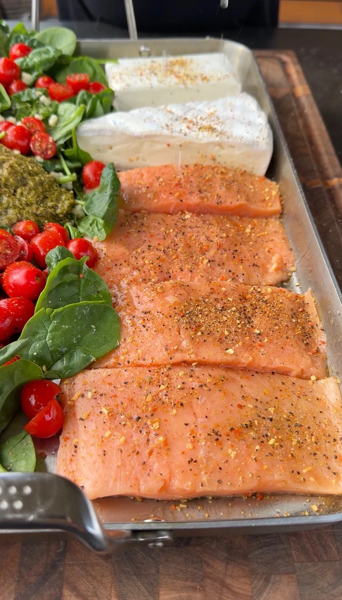 Raw salmon portions seasoned with lemon pepper, Brie, cream cheese, cherry tomatoes, spinach, and pesto in a baking dish
