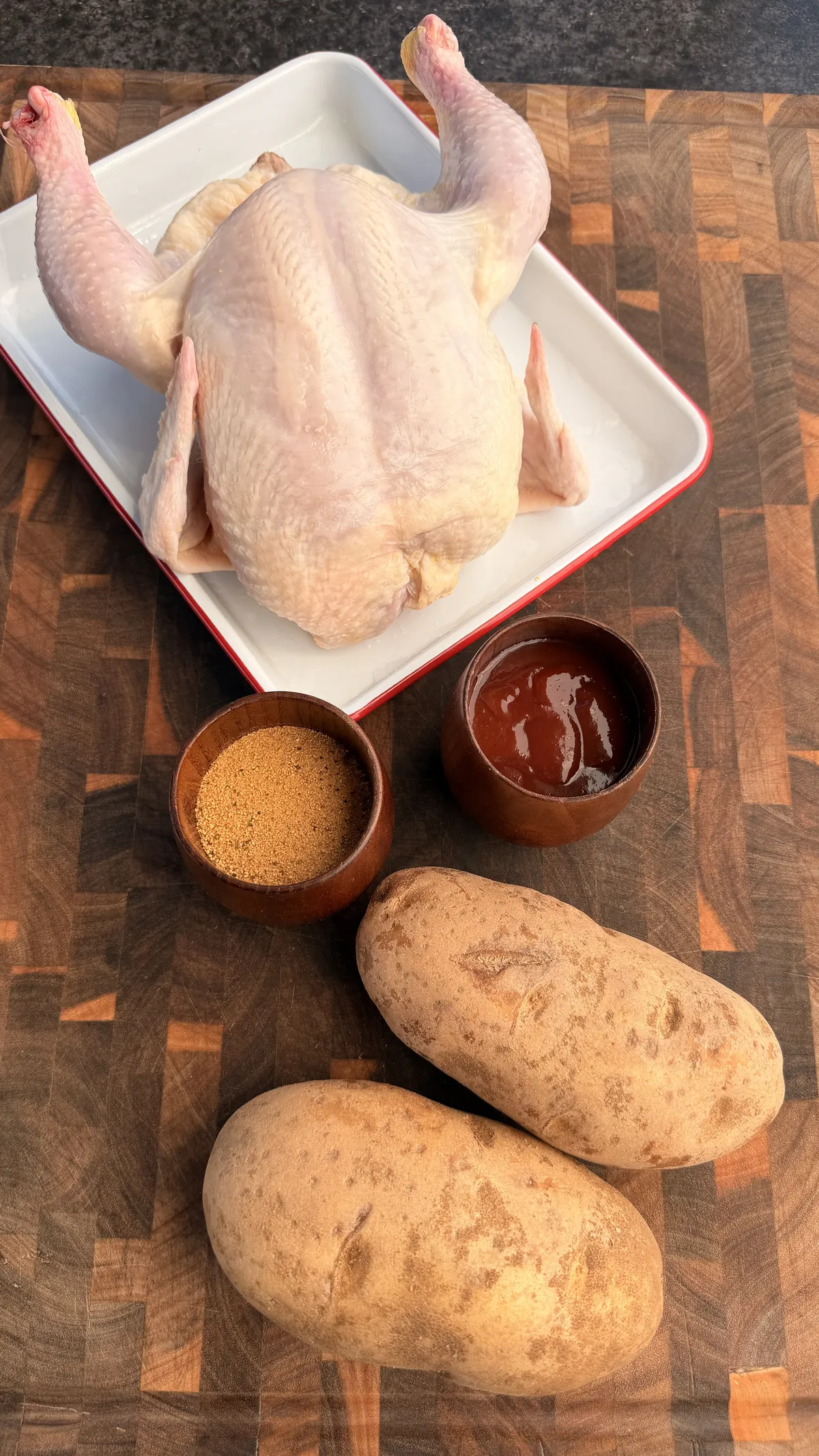 Raw whole chicken in white tray with russet potatoes, poultry seasoning, and BBQ sauce on a wood cutting board
