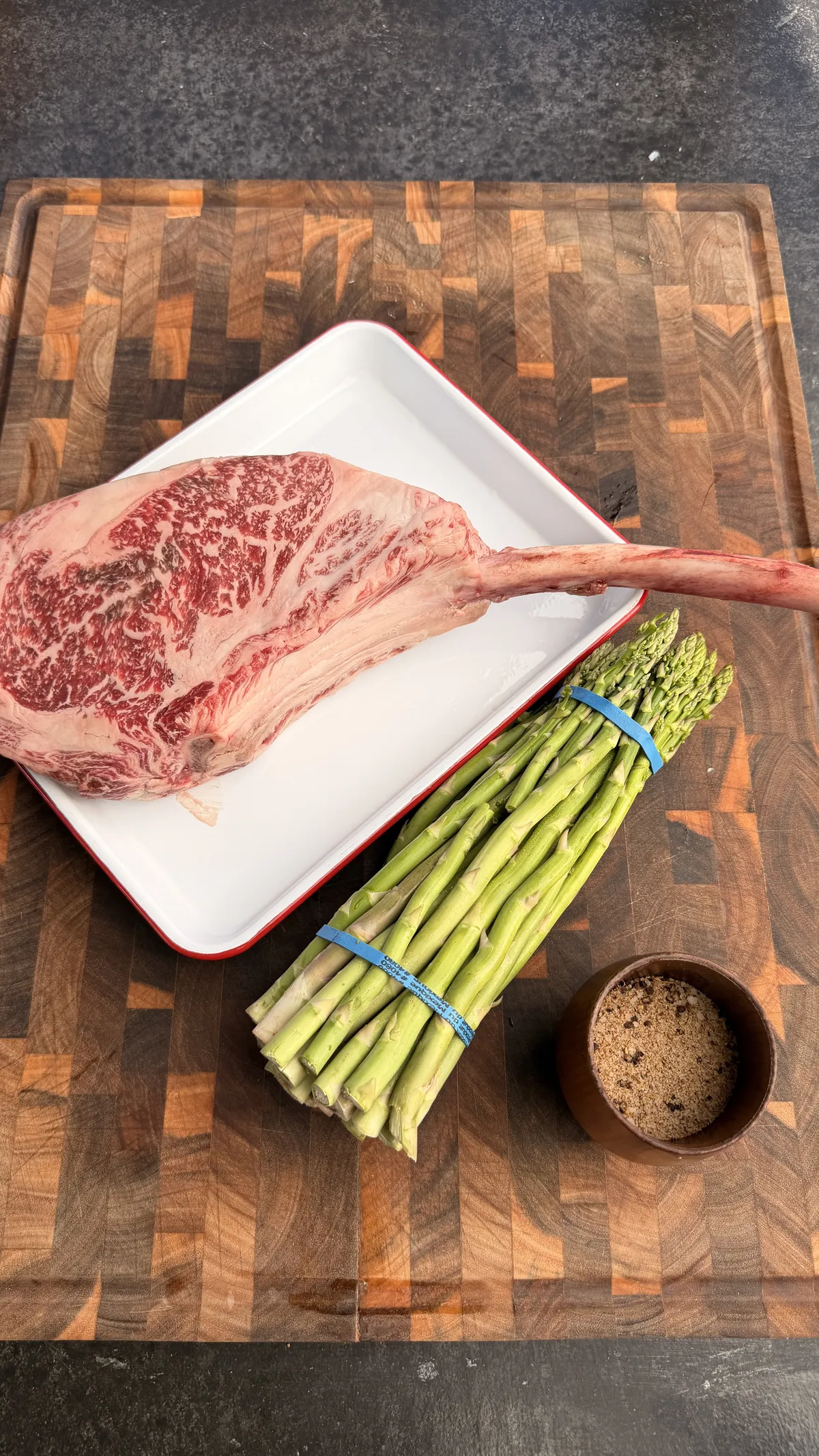 Raw Australian Wagyu tomahawk steak on a white tray with a fresh asparagus bunch and steak seasoning on a wood cutting board