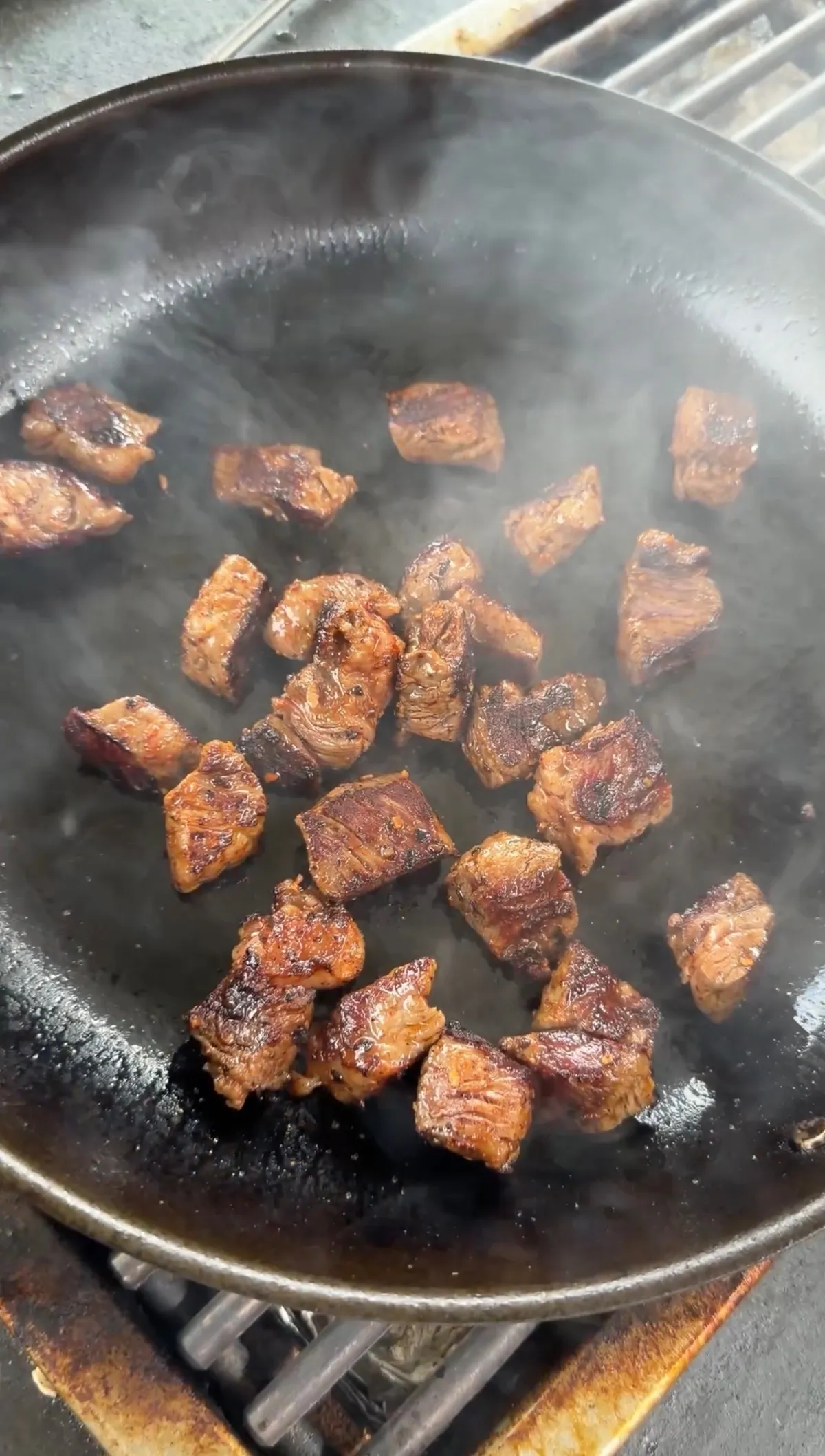 Seasoned ribeye steak bites searing in a hot cast iron skillet with steam rising and golden-brown crust