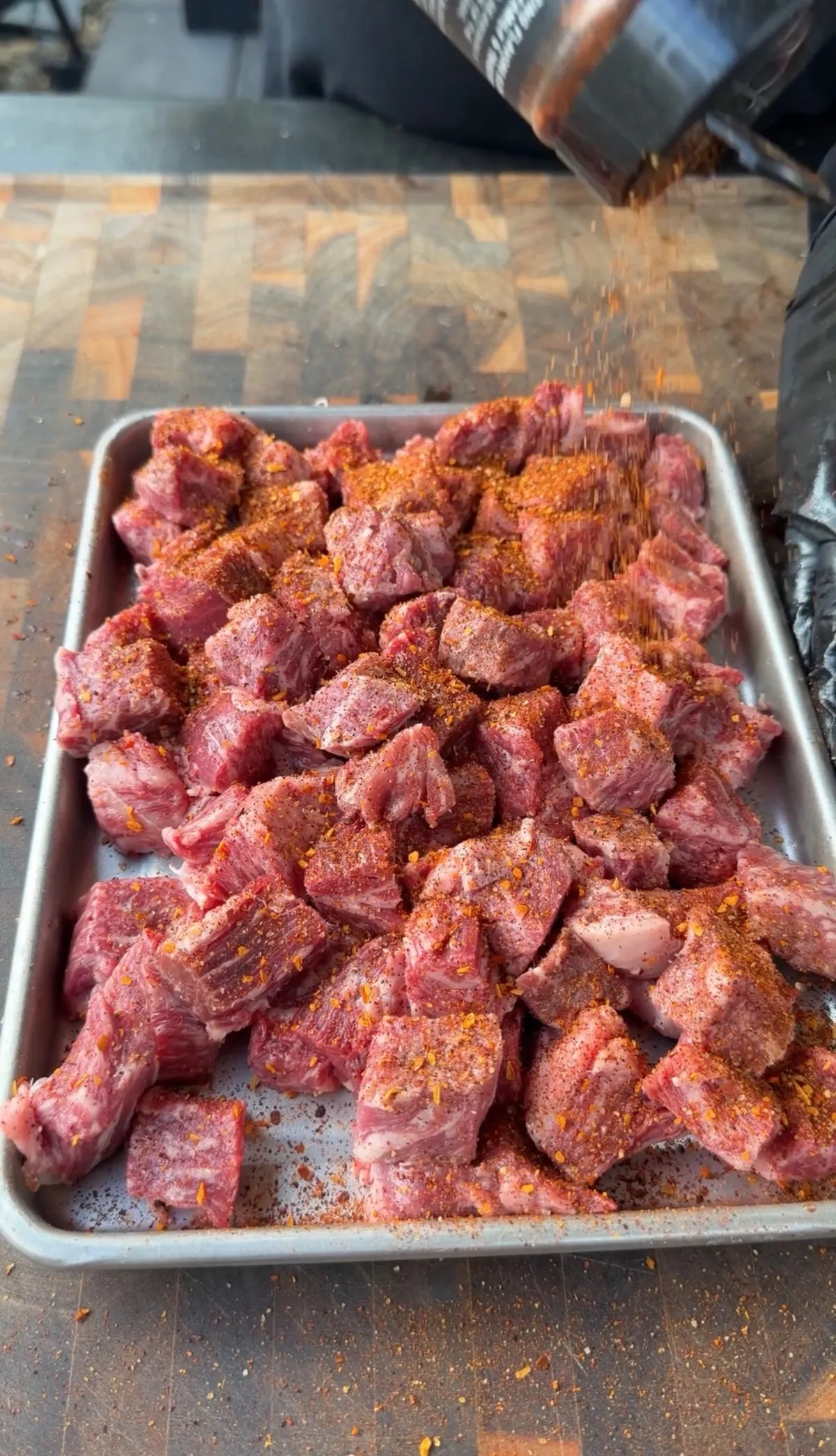 Raw ribeye steak bites on a sheet pan being generously seasoned with steak seasoning before searing