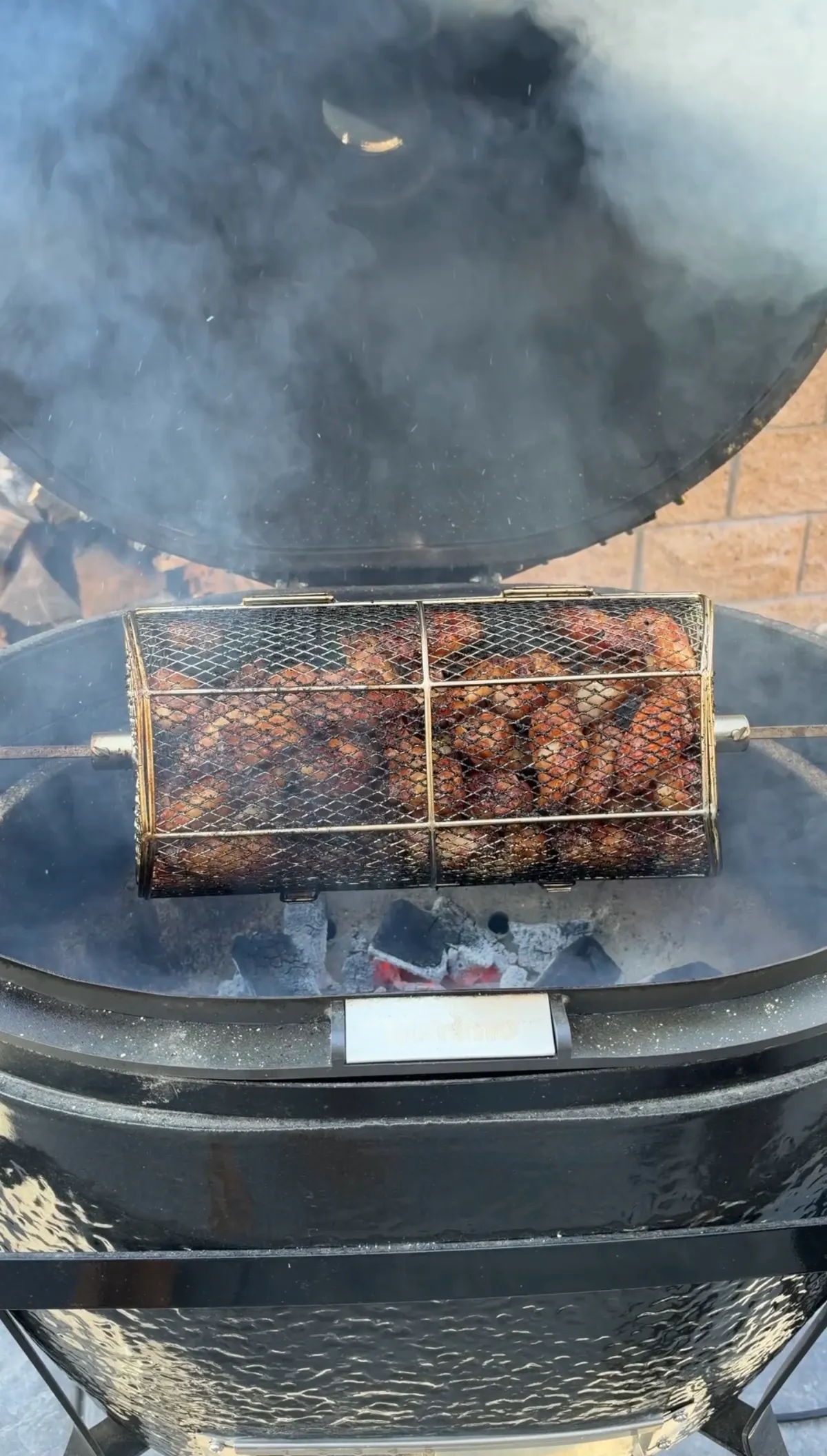 Chicken wings cooking in a rotisserie basket on a charcoal grill with smoke rising, skin turning golden brown and crispy