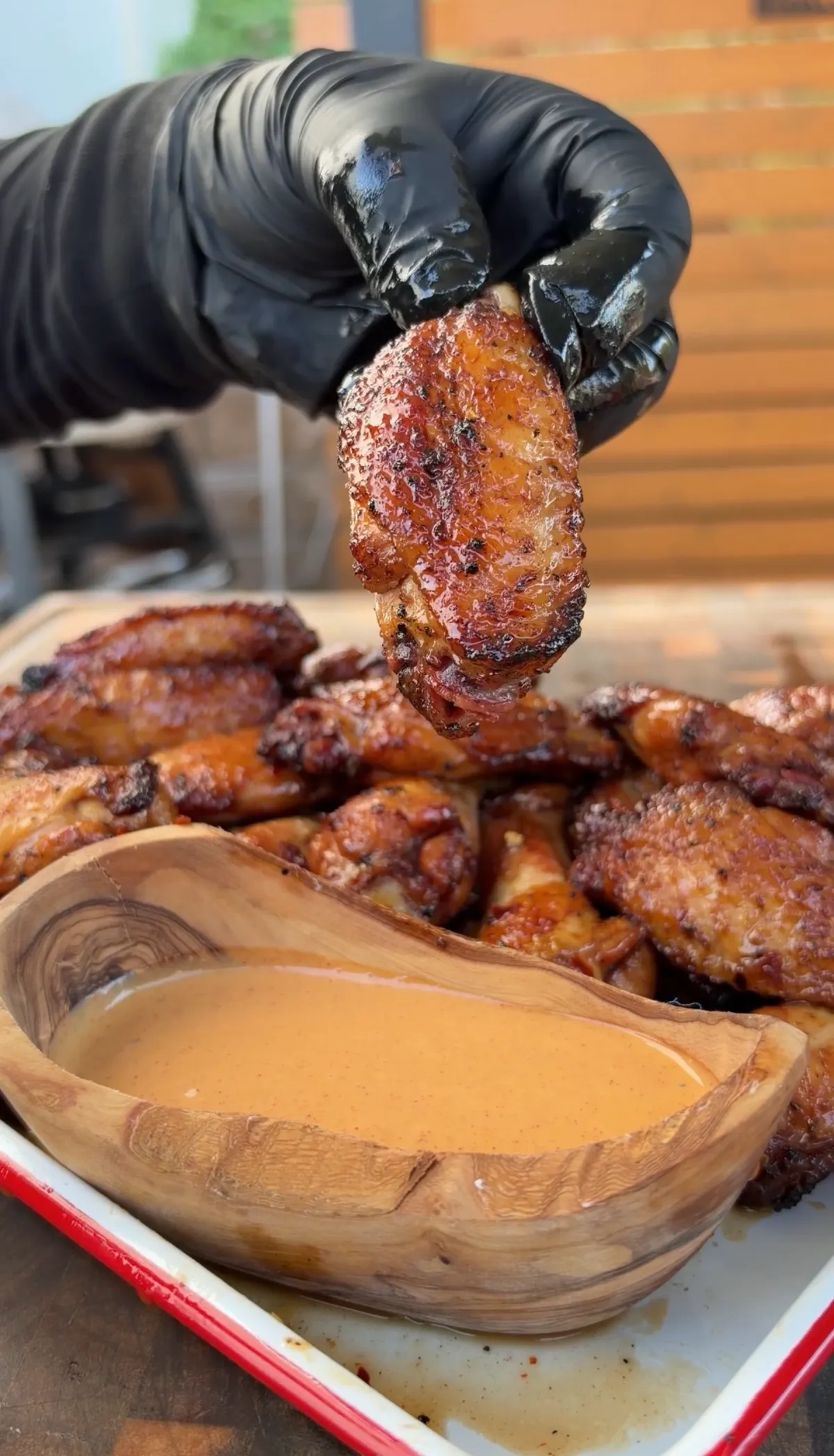 Hand in black glove holding crispy rotisserie chicken wing over hot honey lime dipping sauce in wooden bowl