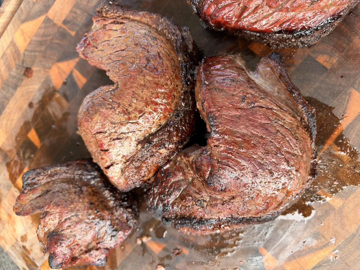 Four folded picanha steaks with dark crust resting on a wooden cutting board after rotisserie cooking