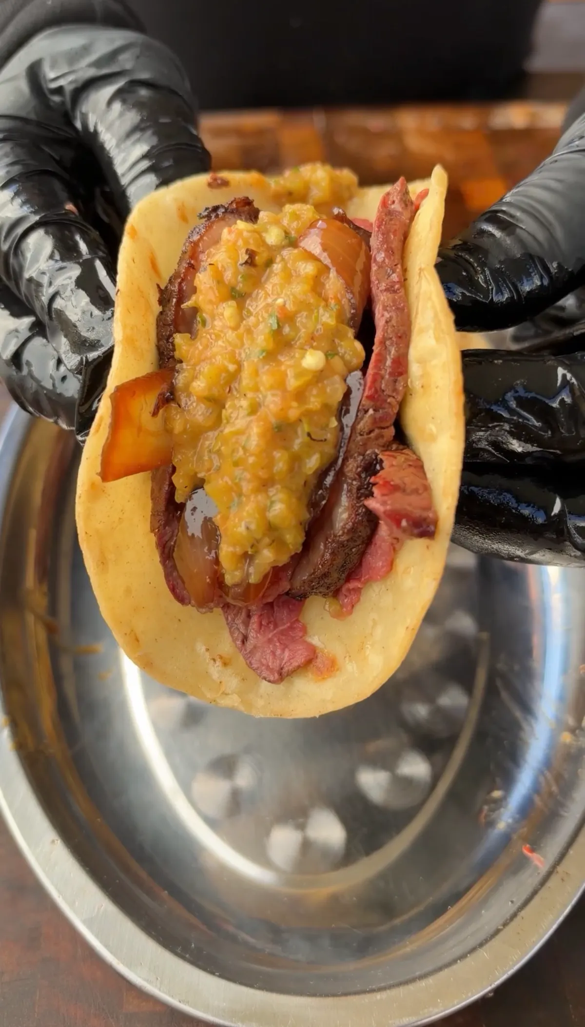 Hands in black gloves holding a corn tortilla filled with thinly sliced picanha, sautéed onion, and roasted Hatch chile salsa